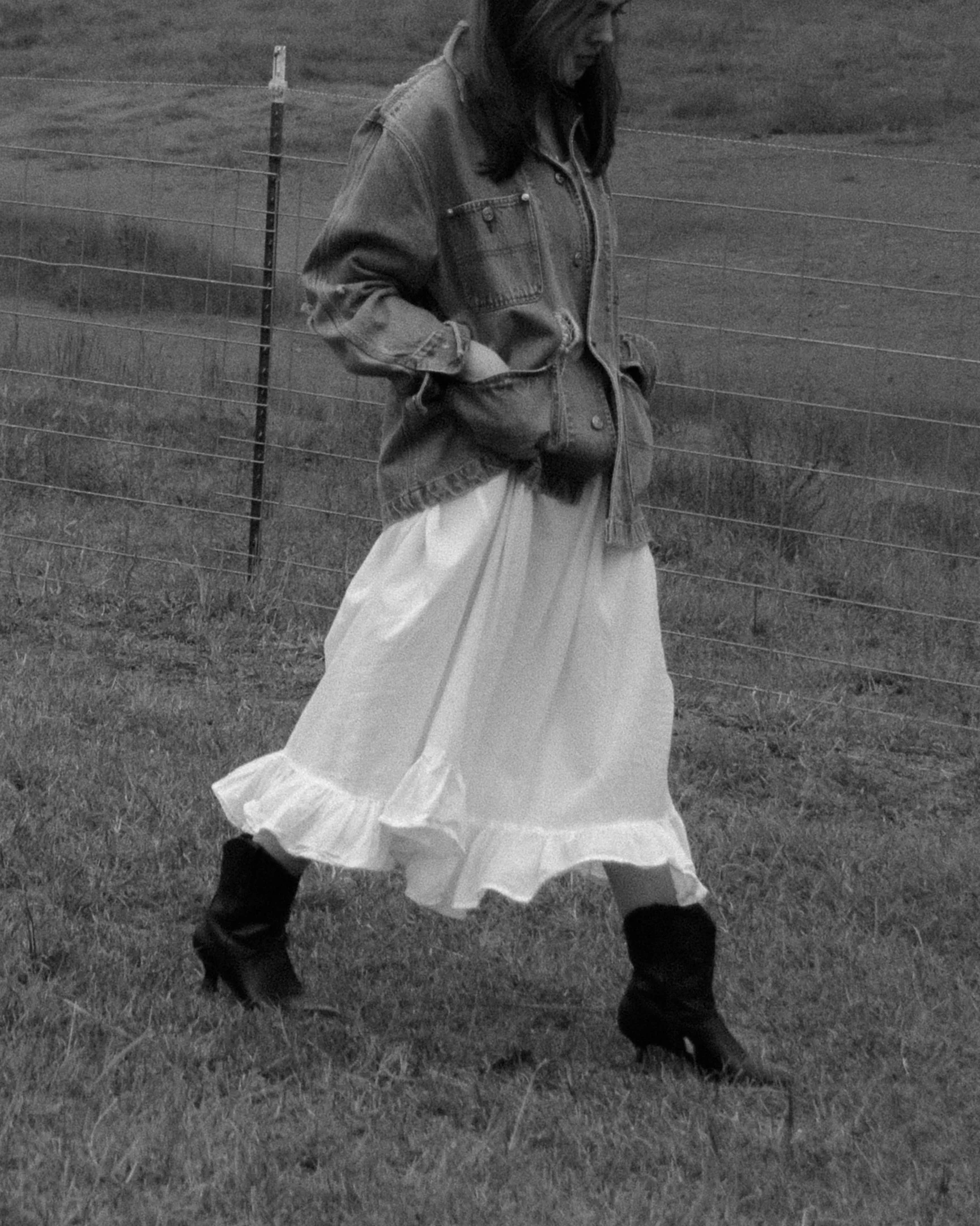 A woman is standing outdoors in front of a wire fence on a grassy field, wearing a denim jacket, a long light-colored skirt, and black boots, with her hands in her pockets.