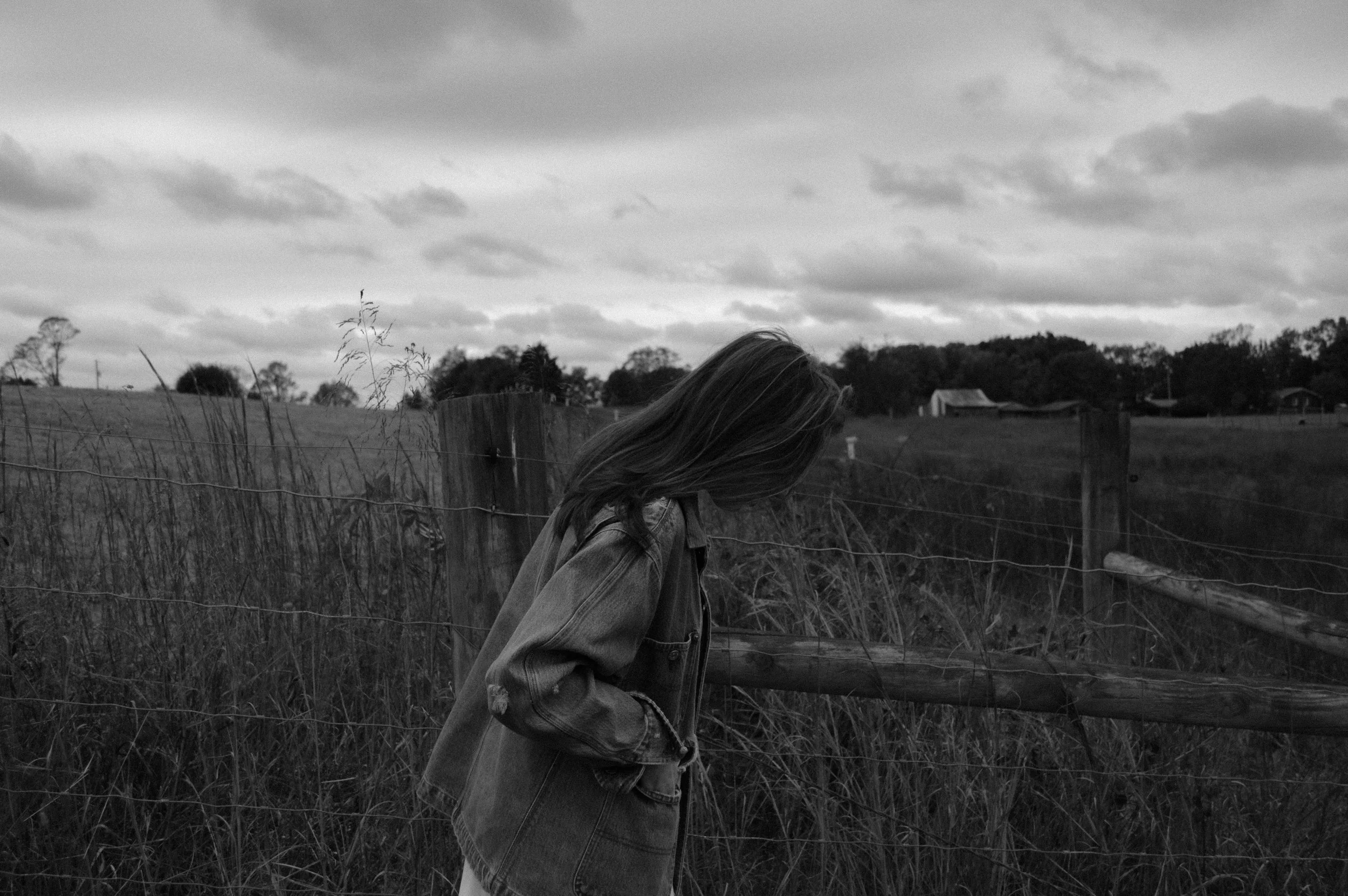 A person with long hair wearing a denim jacket walks along a rural fence at dusk on a cloudy day.