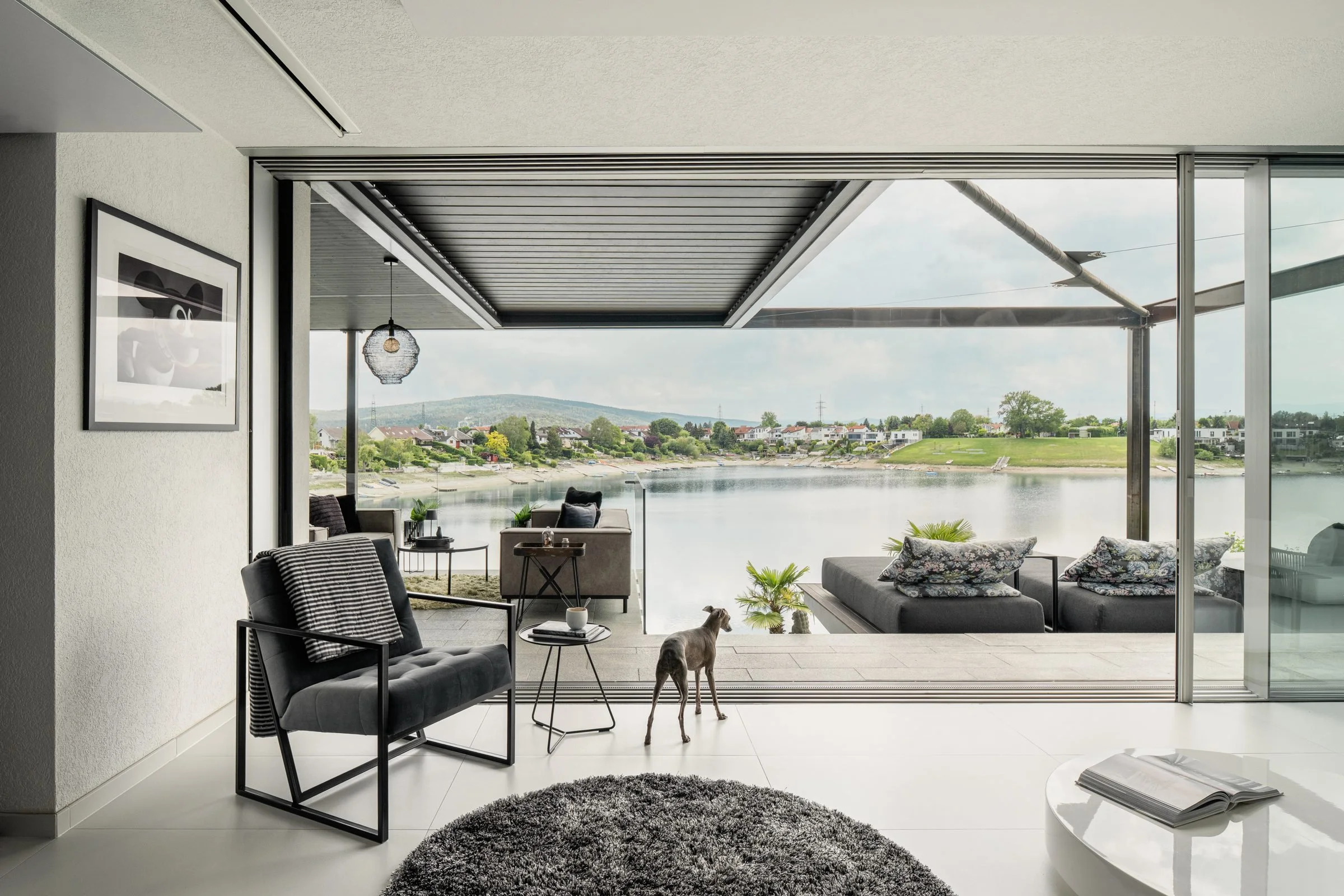 Innenraum eines modernen Wohnzimmers mit großen Glasfenstern, Blick auf einen Fluss und grüne Hügel im Hintergrund, mit schwarzen und grauen Möbeln, Kissen und einer dekorativen Beleuchtung.