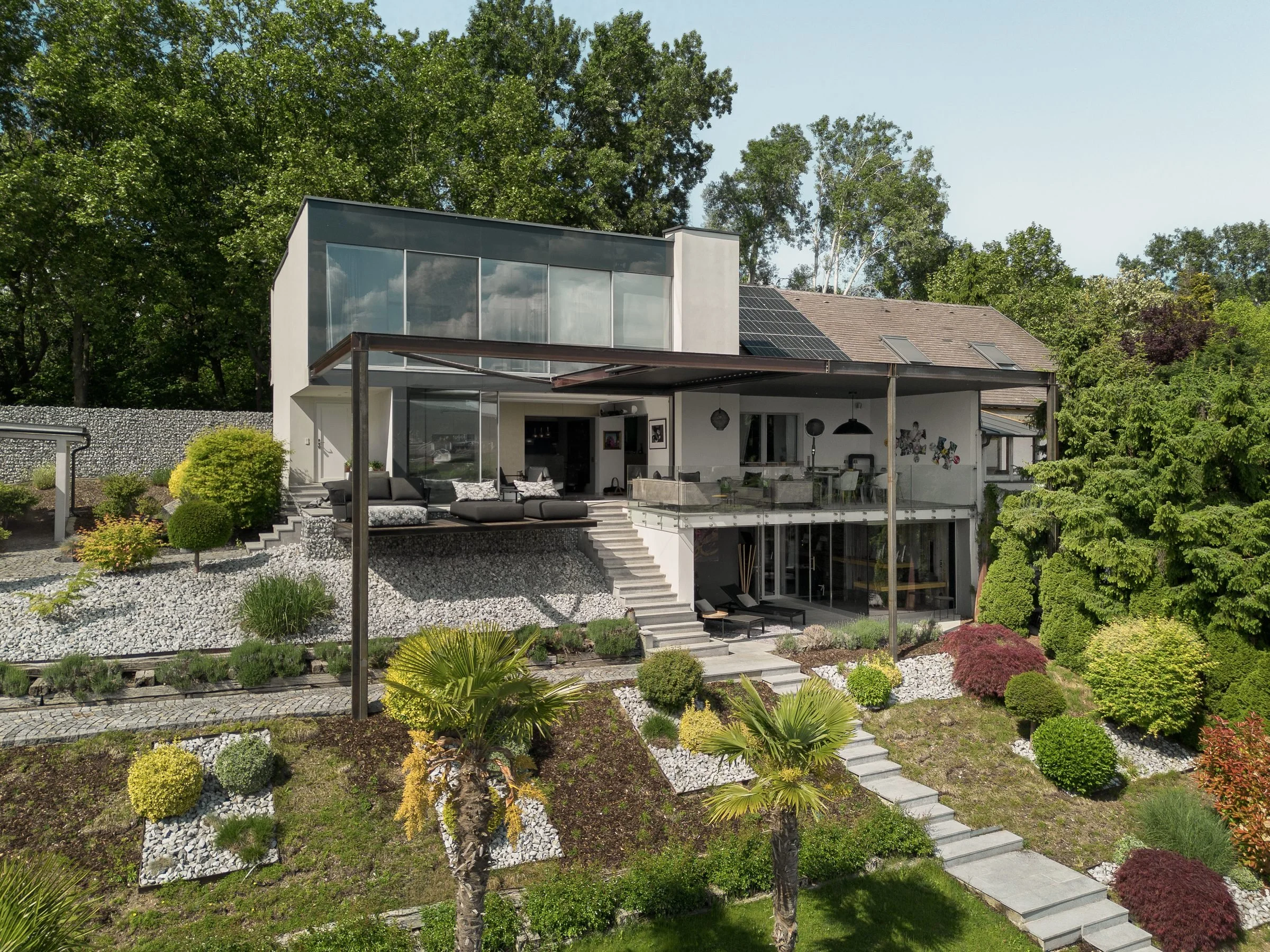 Modernes zweistöckiges Haus mit großen Fenstern, solarpanel auf dem Dach, umgeben von gepflegtem Garten mit Steinen, Bäumen und Sträuchern, sonniger Himmel.