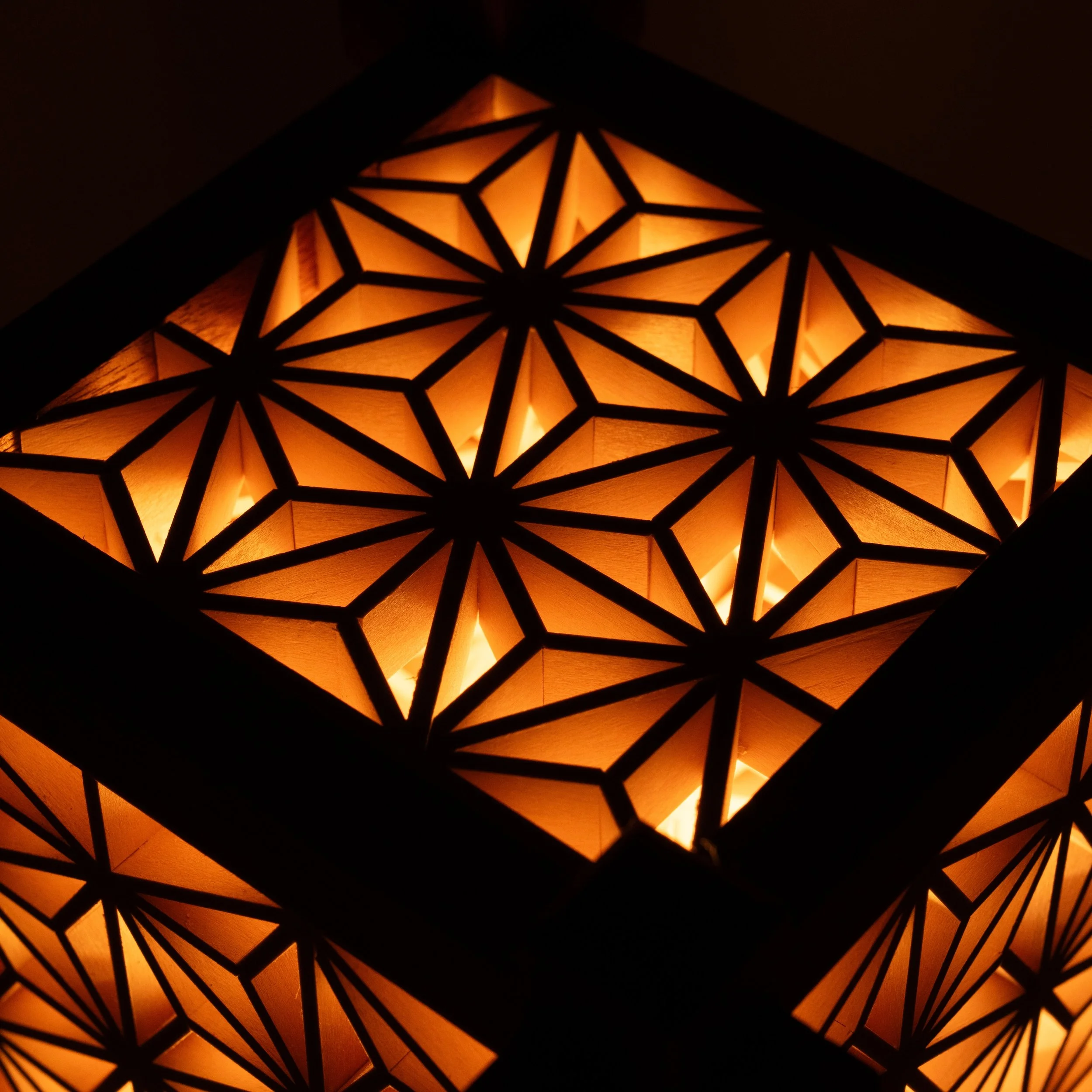 Close-up van het verlichte Asanoha Kumiko-patroon van de handgemaakte Yoake-lamp, met warm oranje licht.