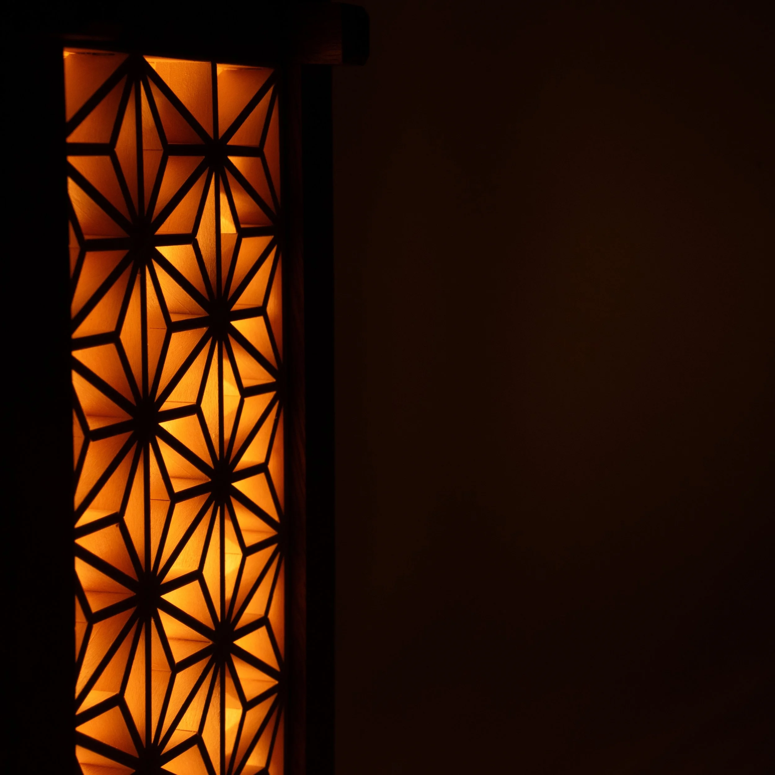 Zijaanzicht van de Yoake-lamp met houten frame en verlichte Kumiko-panelen die warm oranje licht verspreiden.