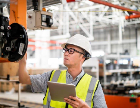 Factory worker wearing a hard hat and safety vest inspecting machinery with a tablet