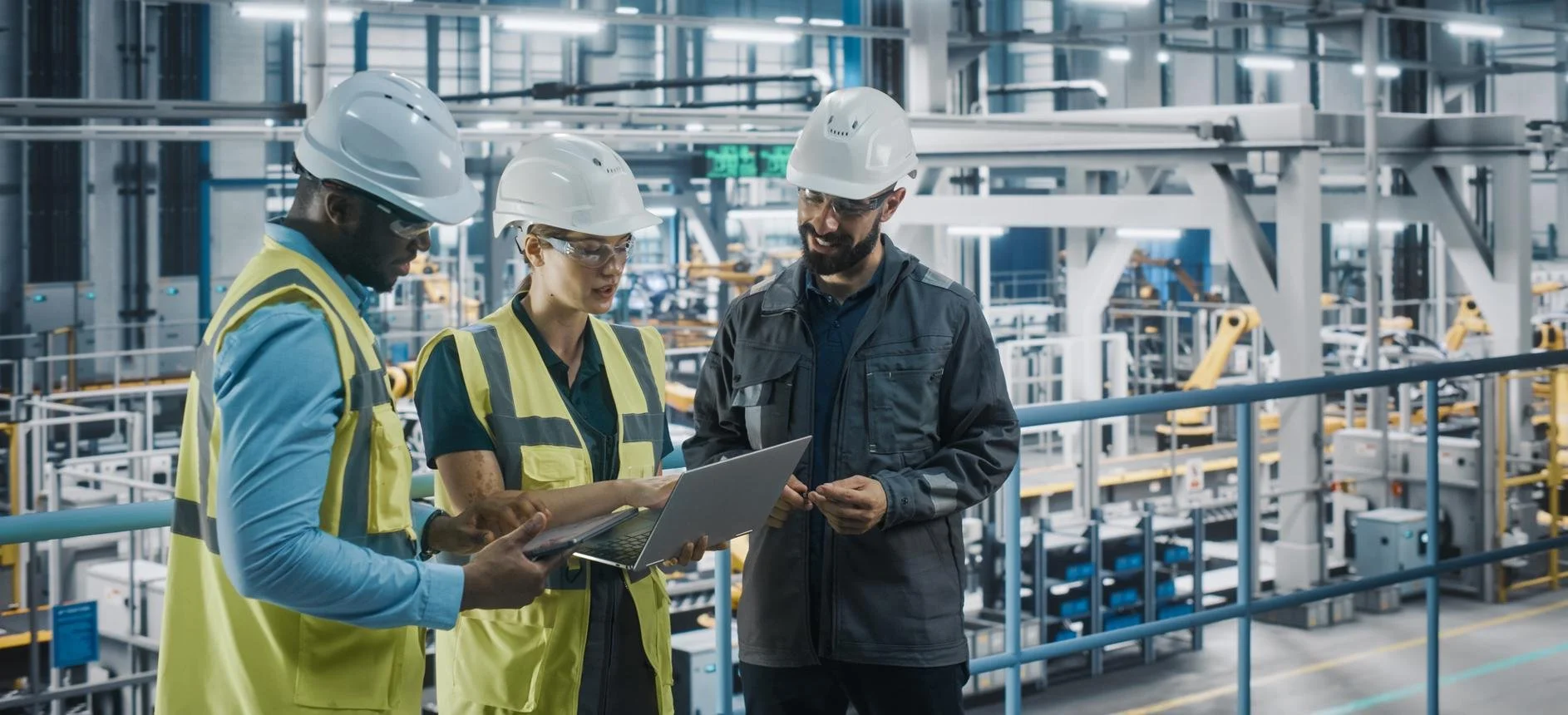 Three industrial workers in hard hats and safety vests discuss a laptop inside a modern manufacturing facility.