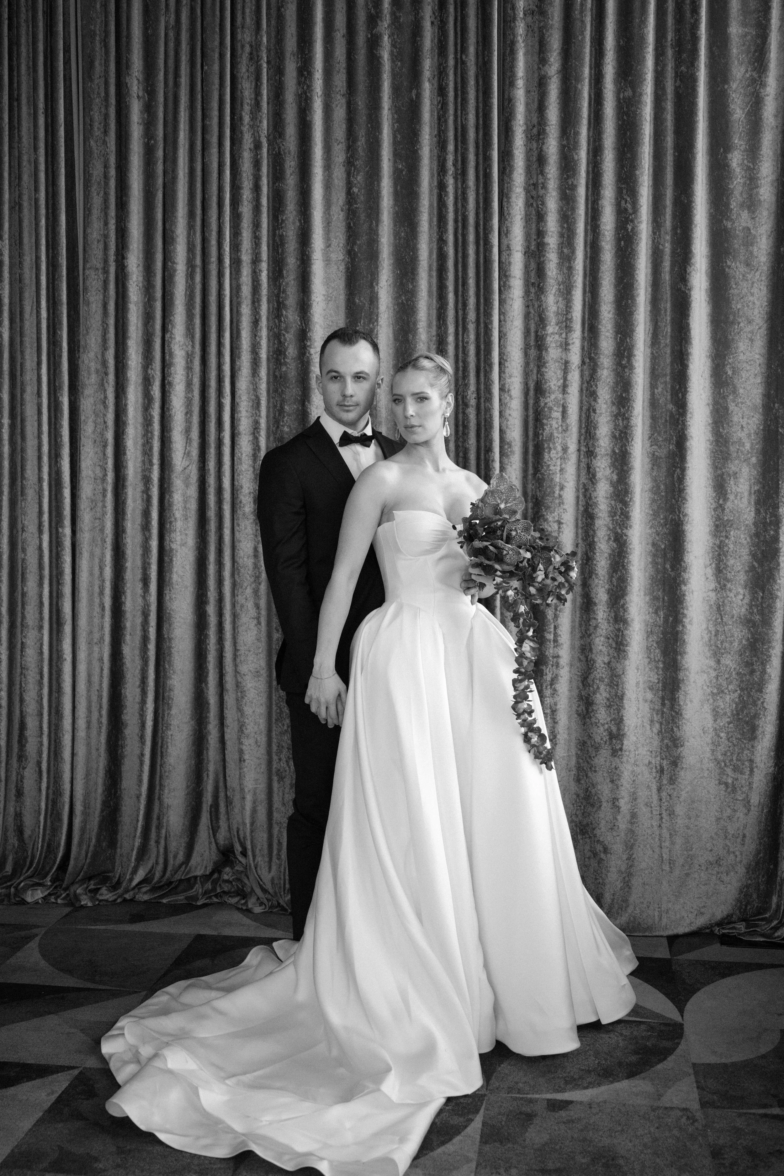Black and white photo of a bride and groom standing close together in front of a curtain backdrop, with the bride holding a bouquet of flowers.