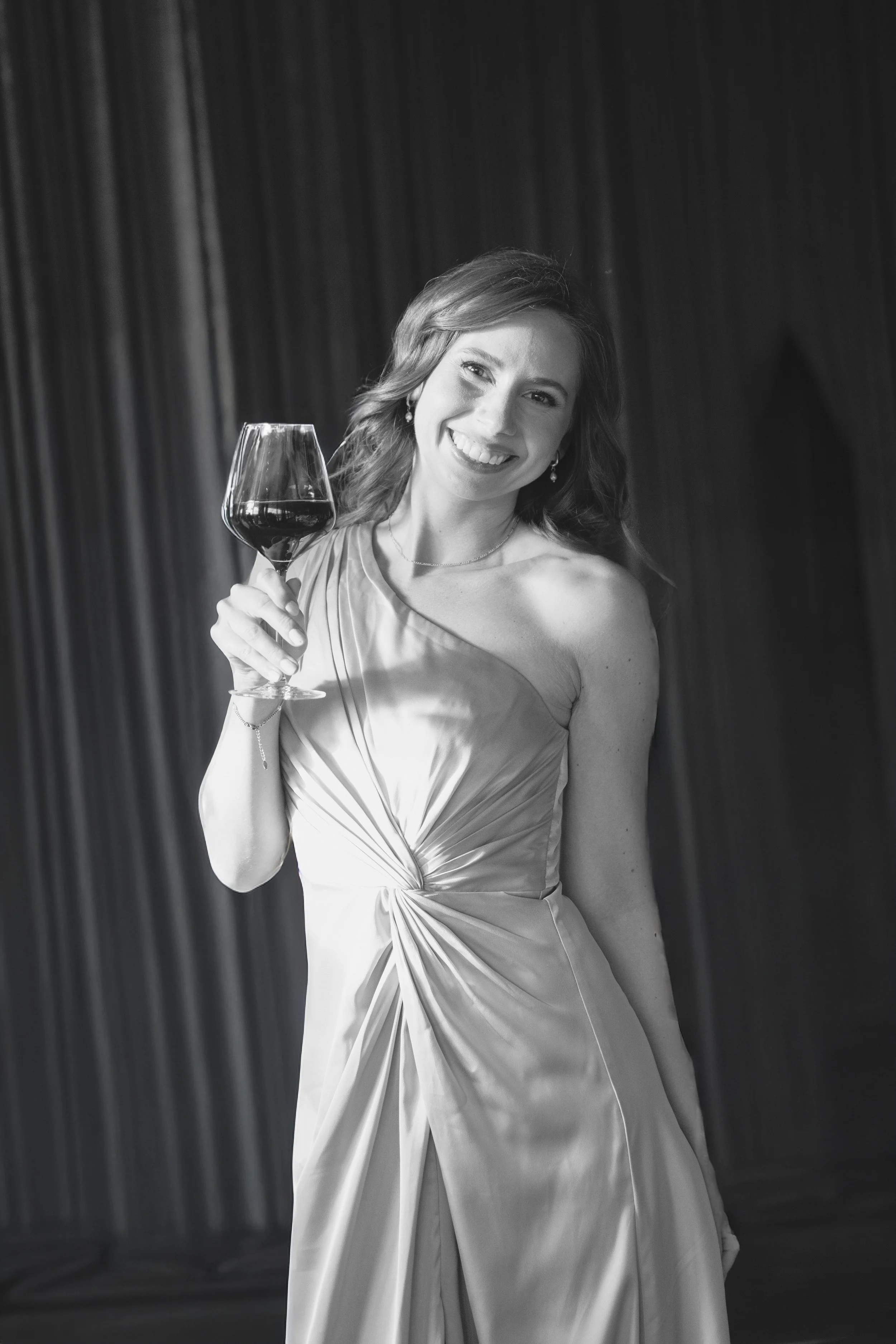 A woman in a elegant dress holding a glass of red wine, smiling, with a dark curtain background.