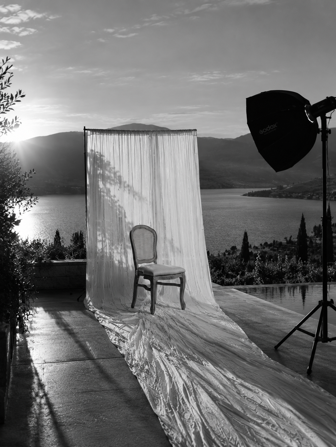 A black-and-white photo of a photography setup with a vintage chair on a fabric-covered platform, set against a scenic Okanagan lake and mountain backdrop at sunset, with studio lighting equipment to the side.