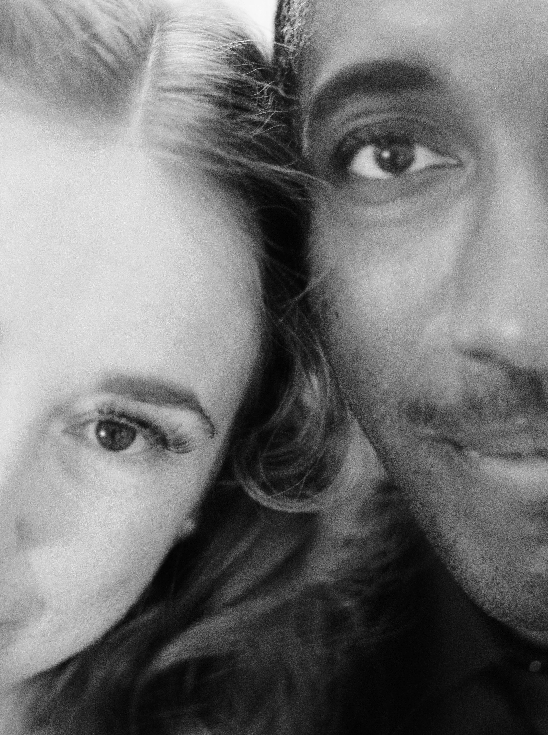 Close-up black and white photo of two people lying with their faces close together, showing eyes and part of their hair.