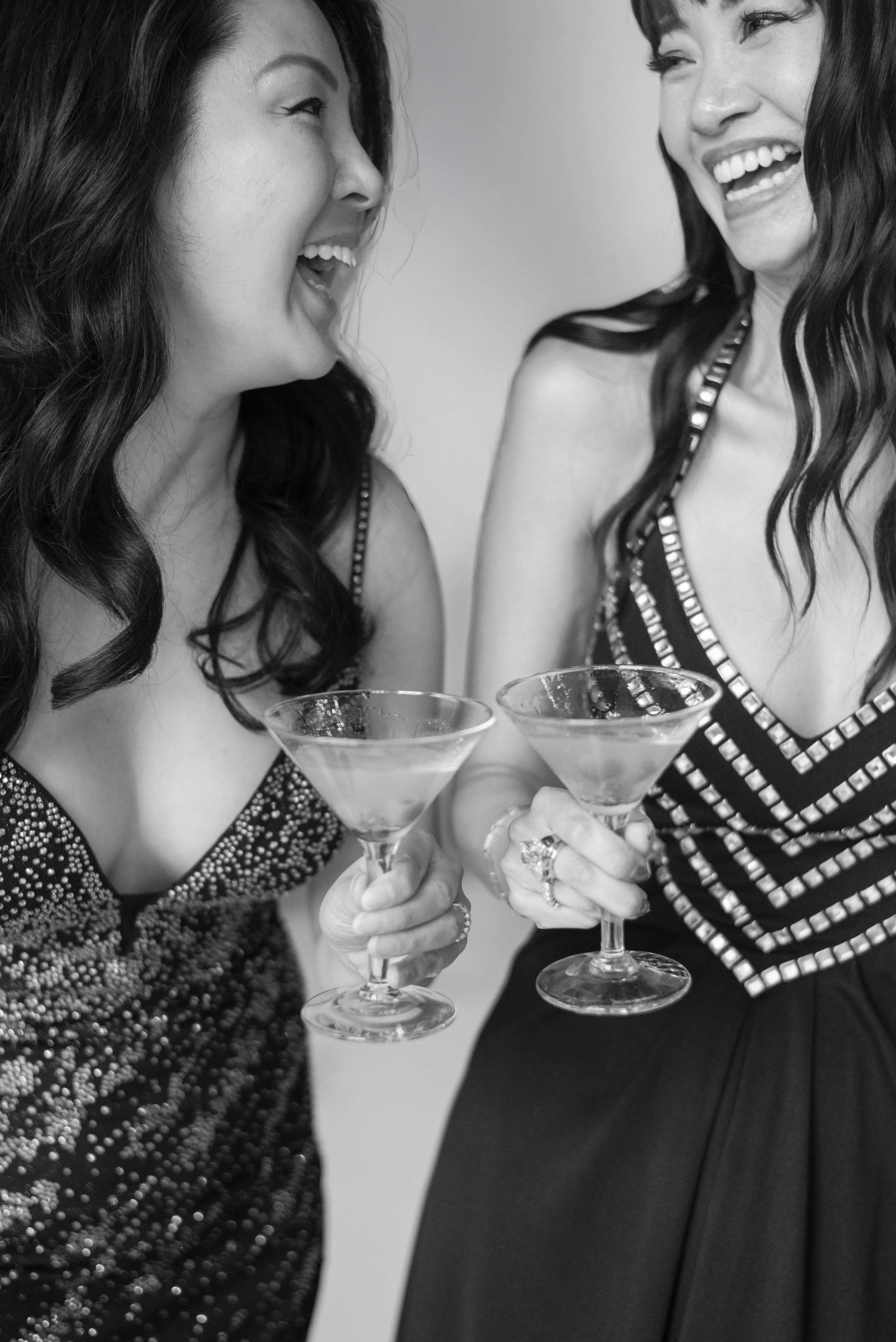 Two women in evening dresses laughing and clinking cocktail glasses at a celebration.