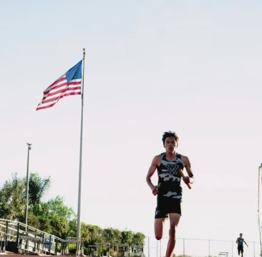A young male athlete running on a track during daytime with an American flag on a flagpole in the background.