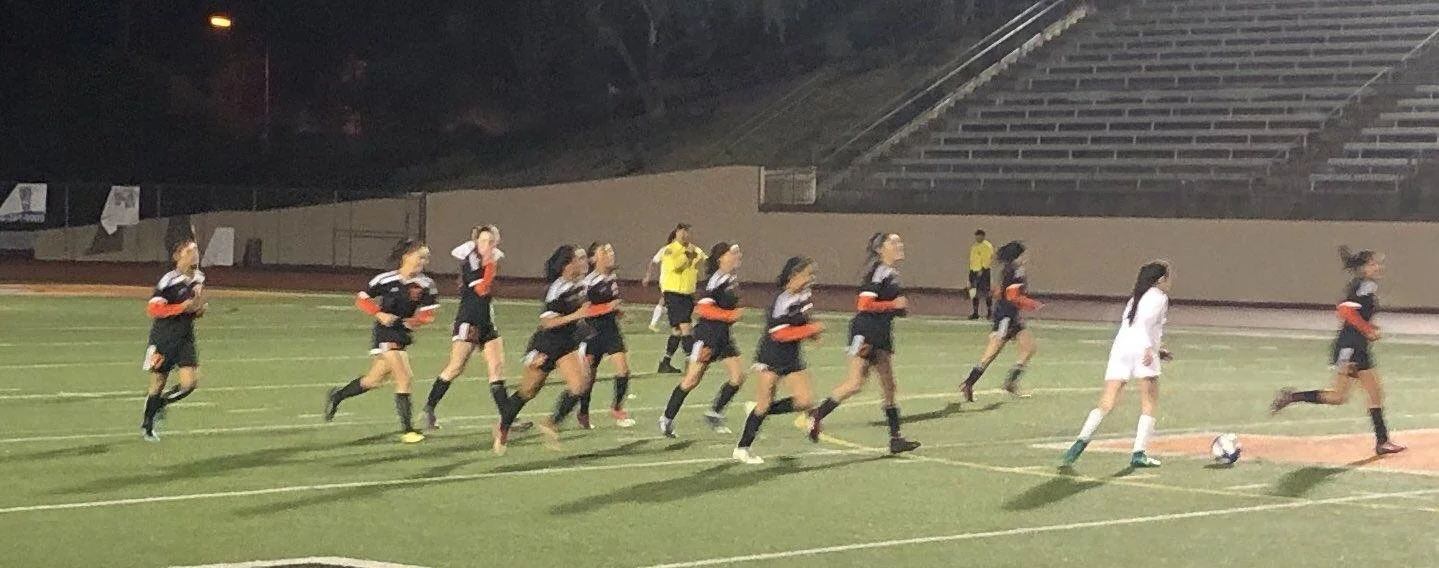 A women's soccer team warming up on a soccer field at night, with a player in white and green preparing to kick the ball as teammates run behind her.