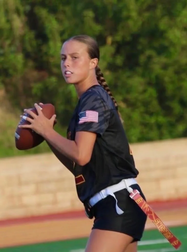 Young woman in an American flag-themed sports uniform holding a football on a football field.