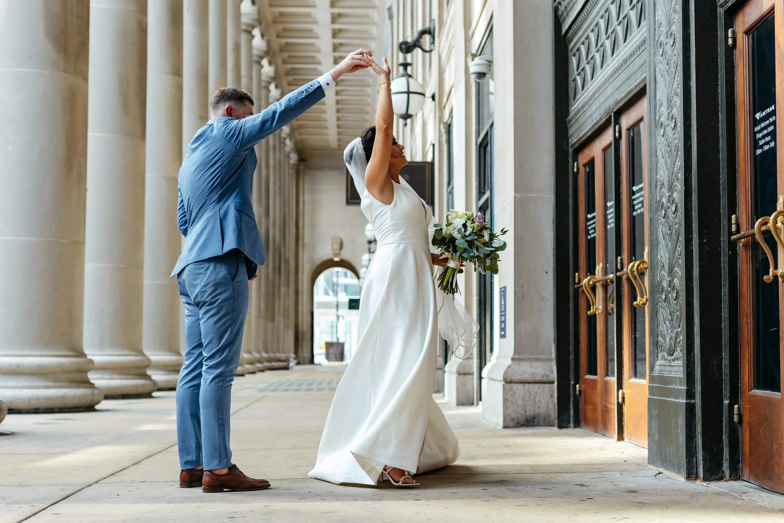 Groom in a blue suit leading his bride through a turn outside a stone building