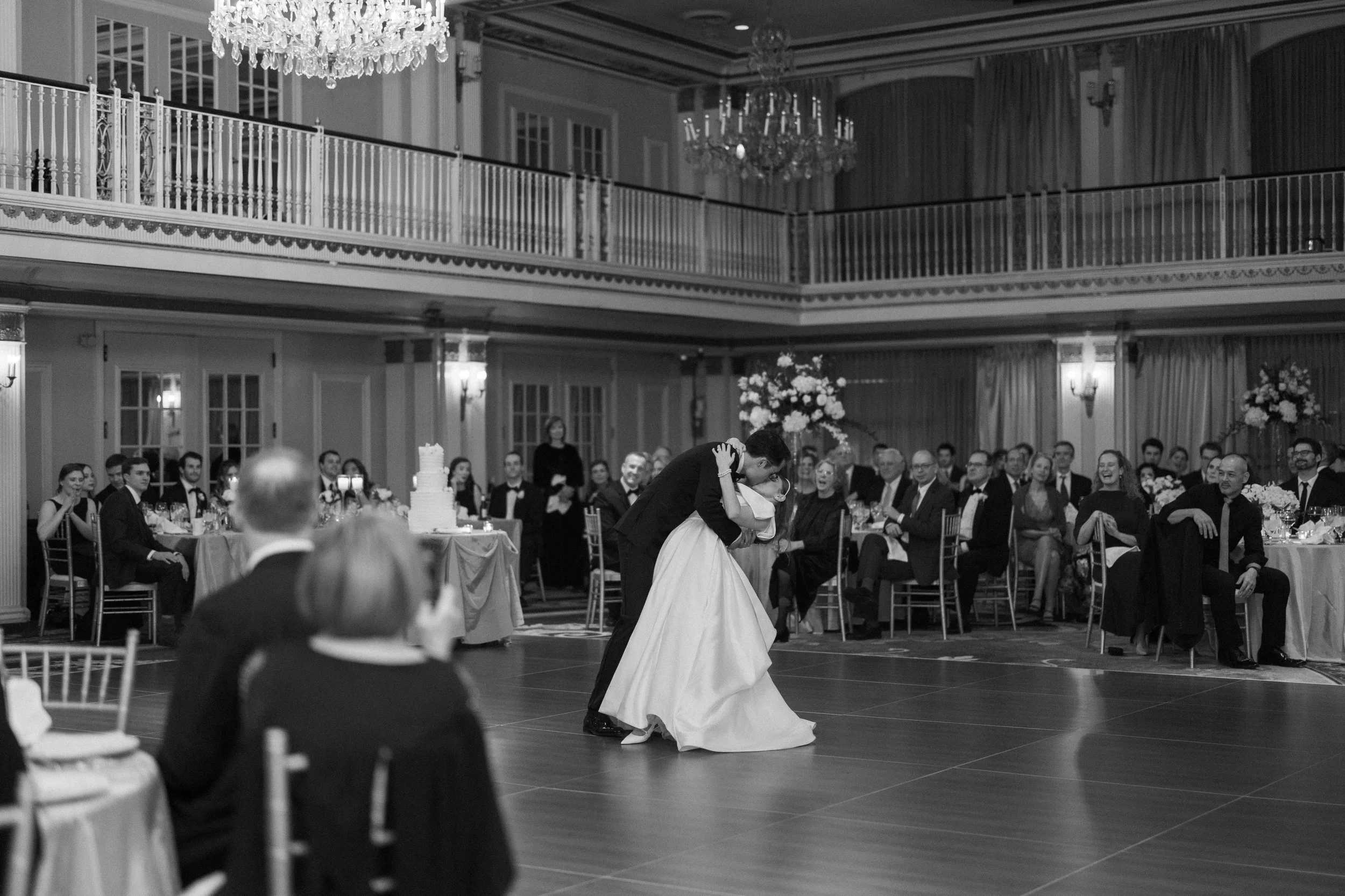Black-and-white photograph of a bride and groom dancing in a grand ballroom surrounded by guests