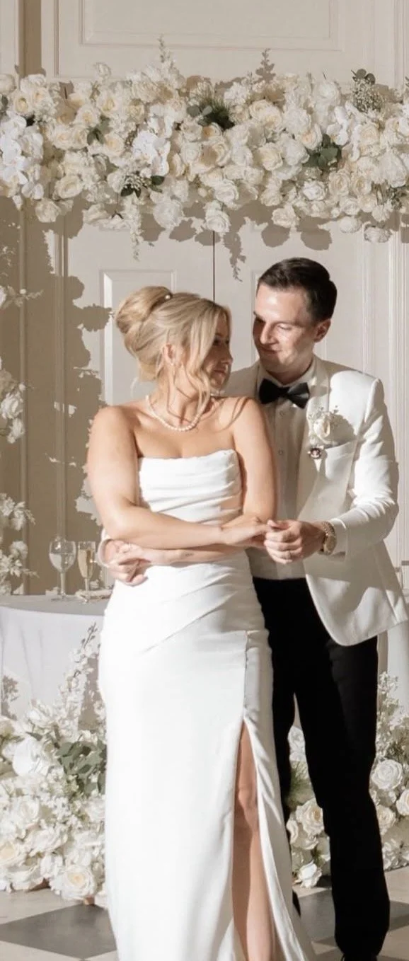 Bride and groom holding hands during their first dance in front of a white floral backdrop