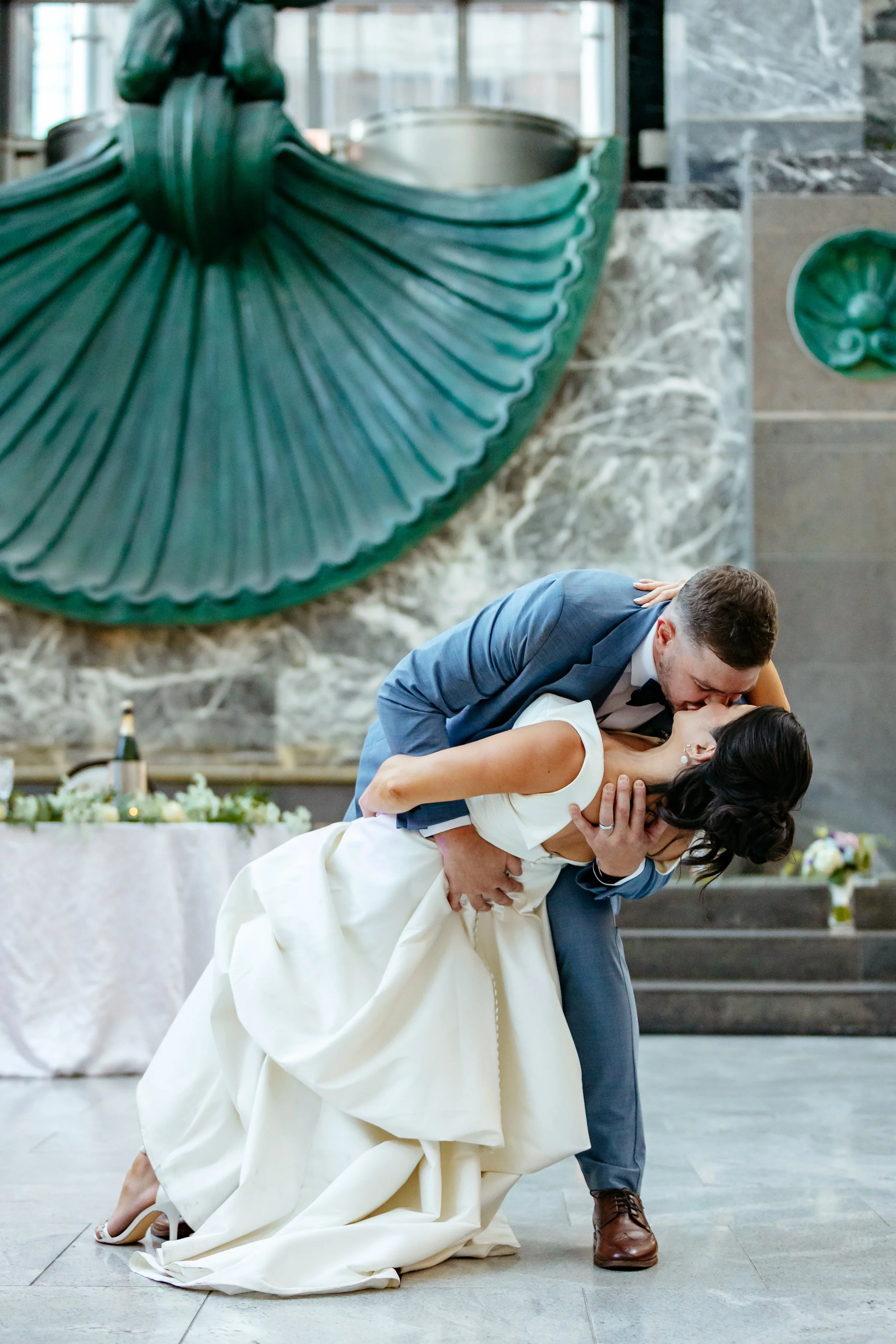 Groom in a blue suit dipping and kissing his bride beside an ornate green staircase