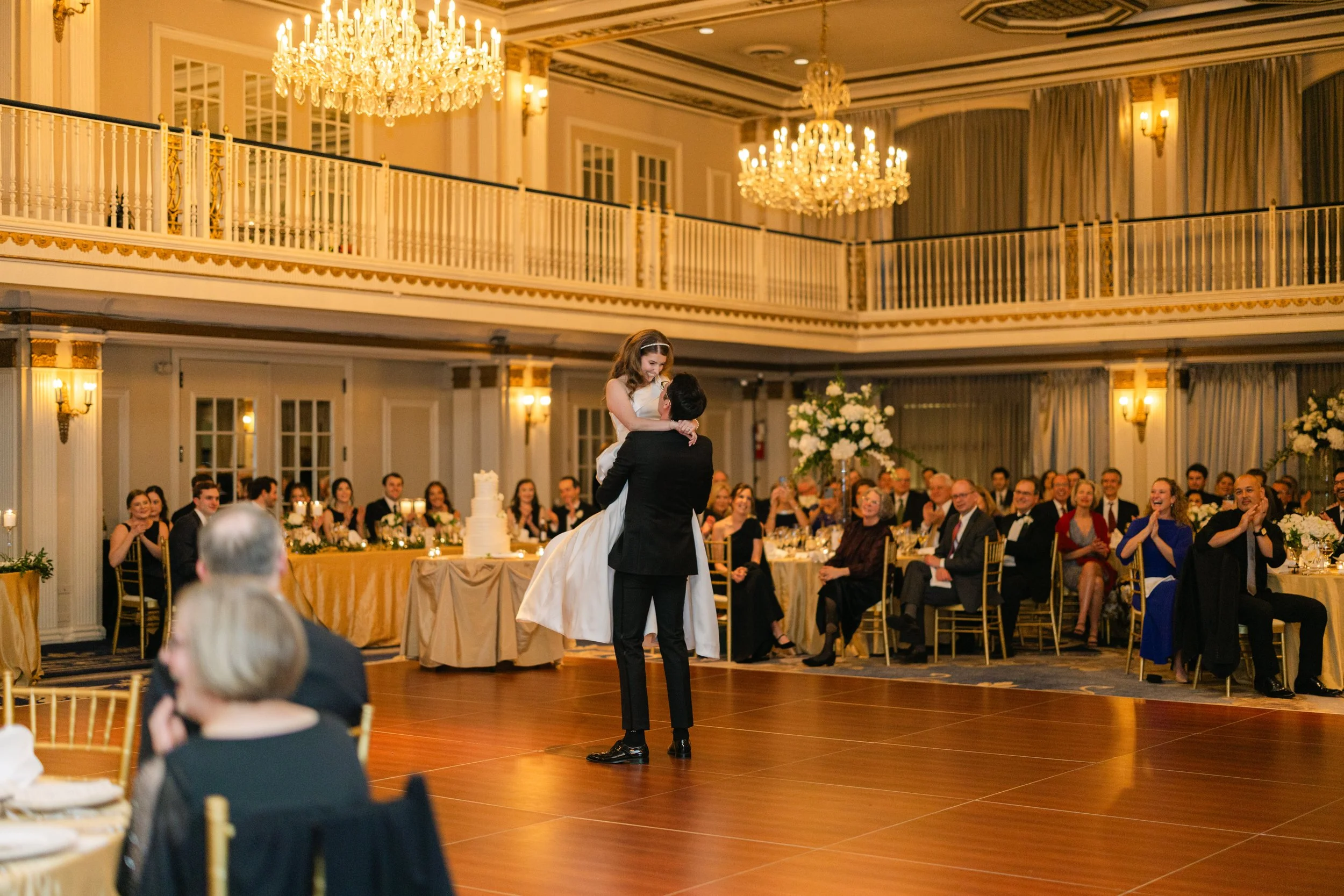 Groom lifting his bride during their first dance in a chandelier-lit ballroom surrounded by guests