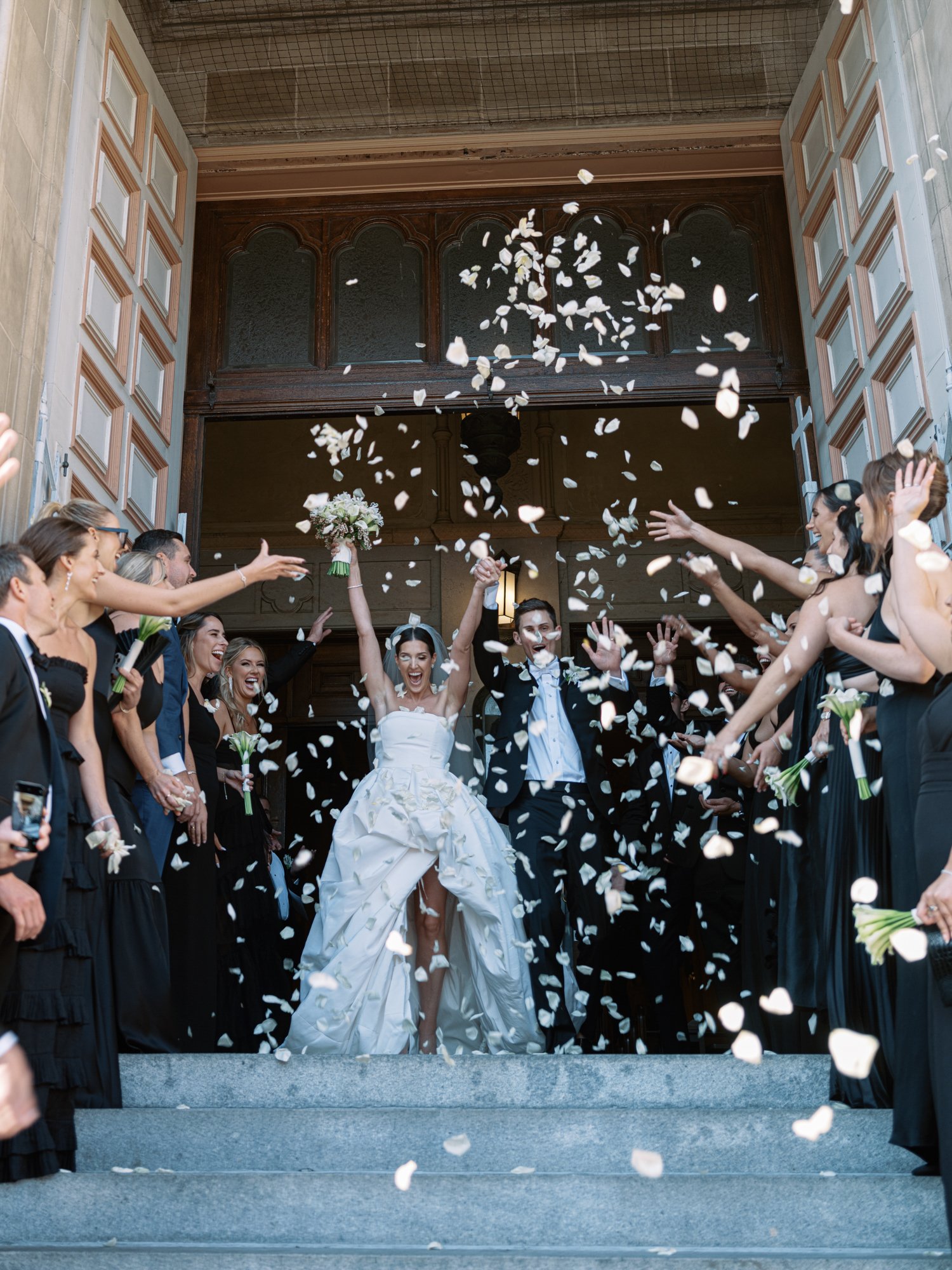 A bride and groom celebrating on wedding day, surrounded by guests throwing flower petals or confetti outside a building entrance.