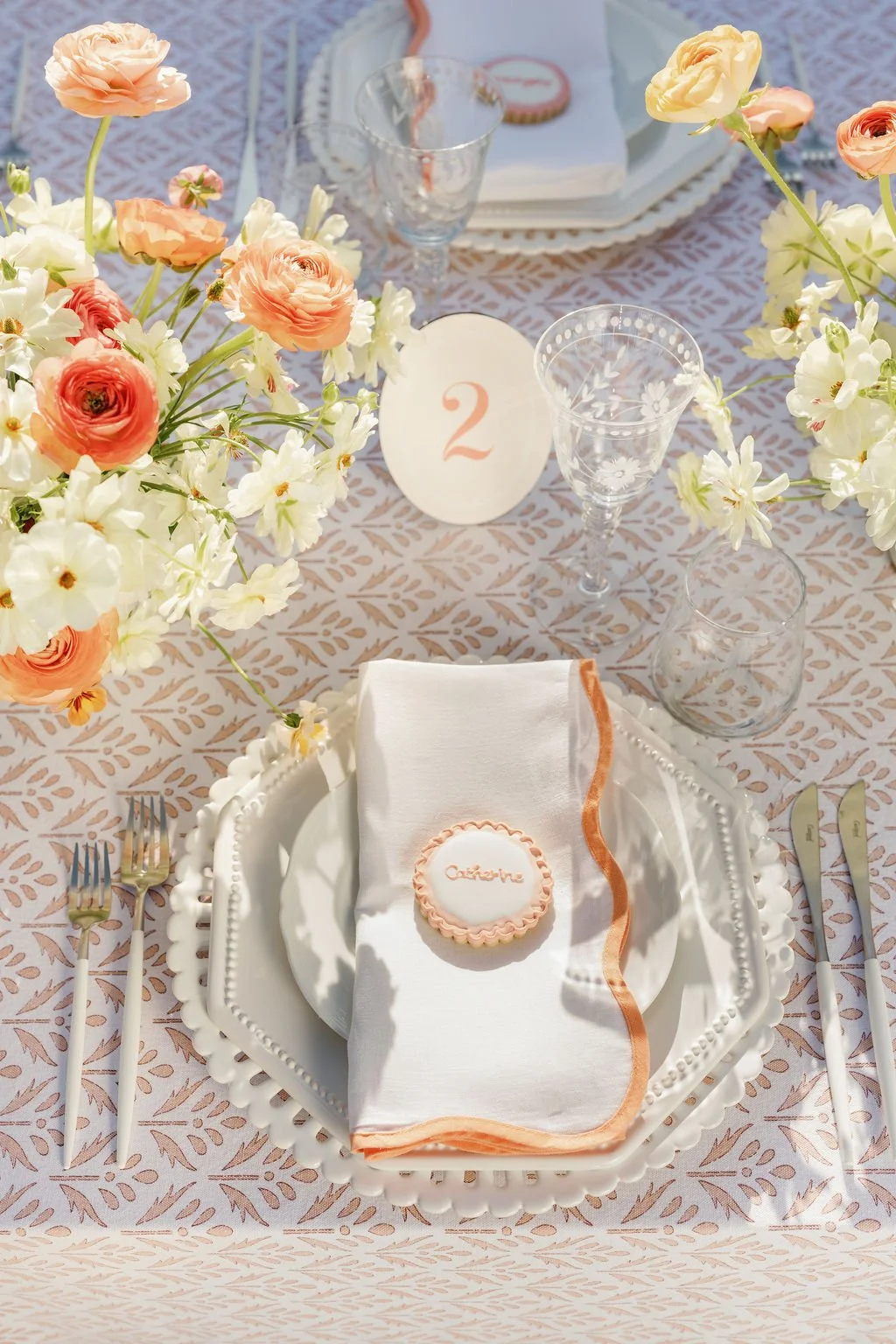A beautifully arranged table for a special event with a white and peach color theme. It includes a white decorative plate with a napkin and a cookie labeled 'Céline', surrounded by flowers in white and peach tones, crystal glasses, and silverware. A small table number card with the number 2 is also present.