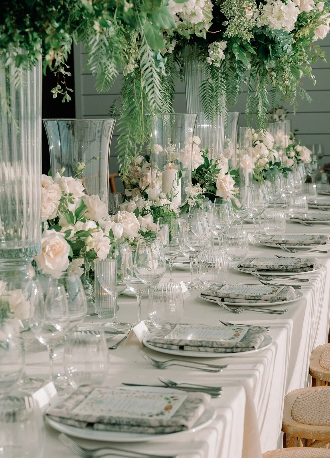 Elegant dining table decorated with white roses and greenery, set with glassware, plates, and silverware for a wedding or special event.