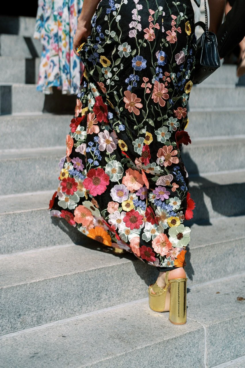Close-up of a person ascending outdoor concrete stairs, wearing a black floral embroidered dress with colorful flowers, gold high-heeled sandals, and carrying a black handbag.