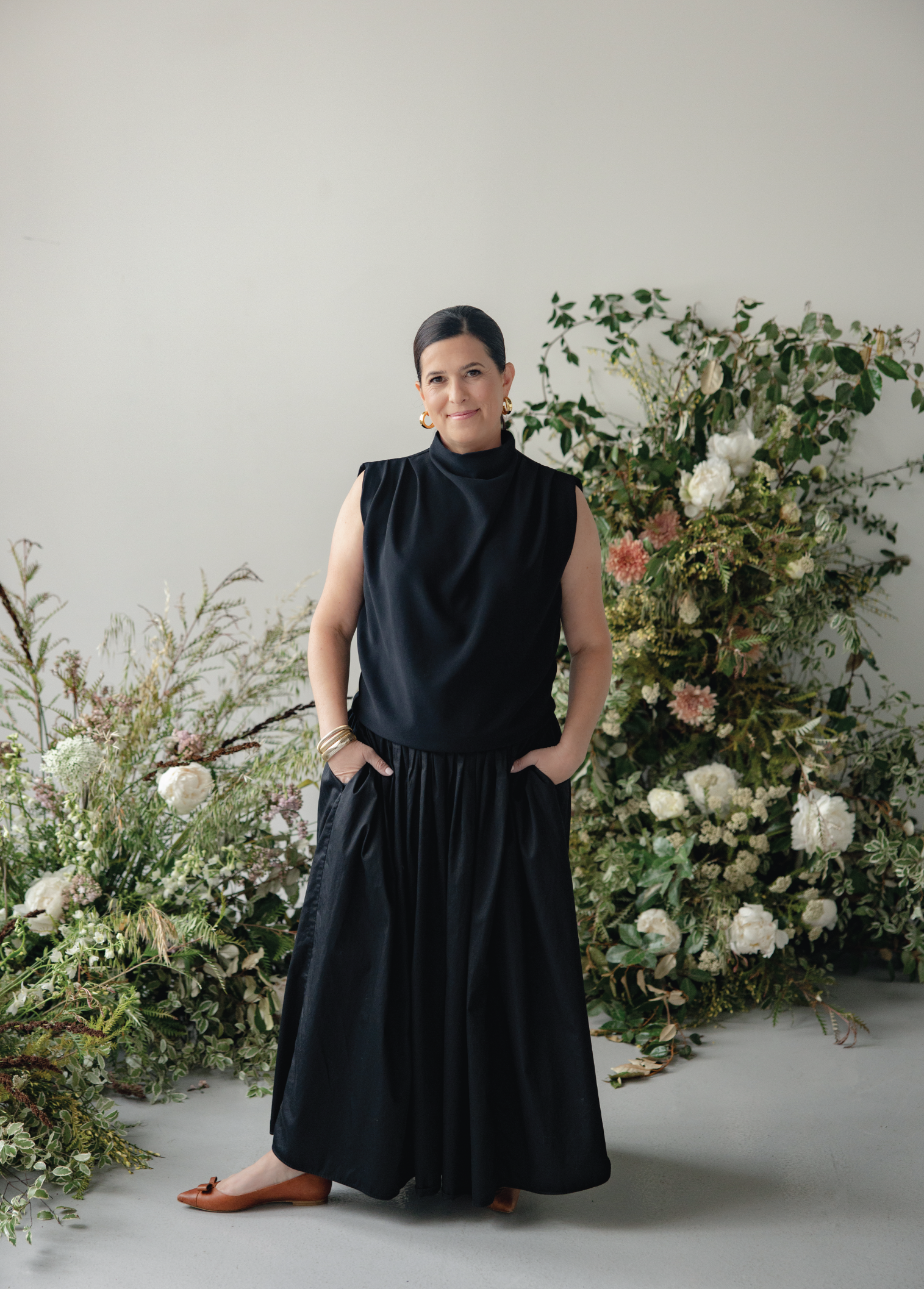 Woman in black sleeveless top and long skirt standing in front of a floral arrangement, with her hands in her pockets and smiling at the camera.