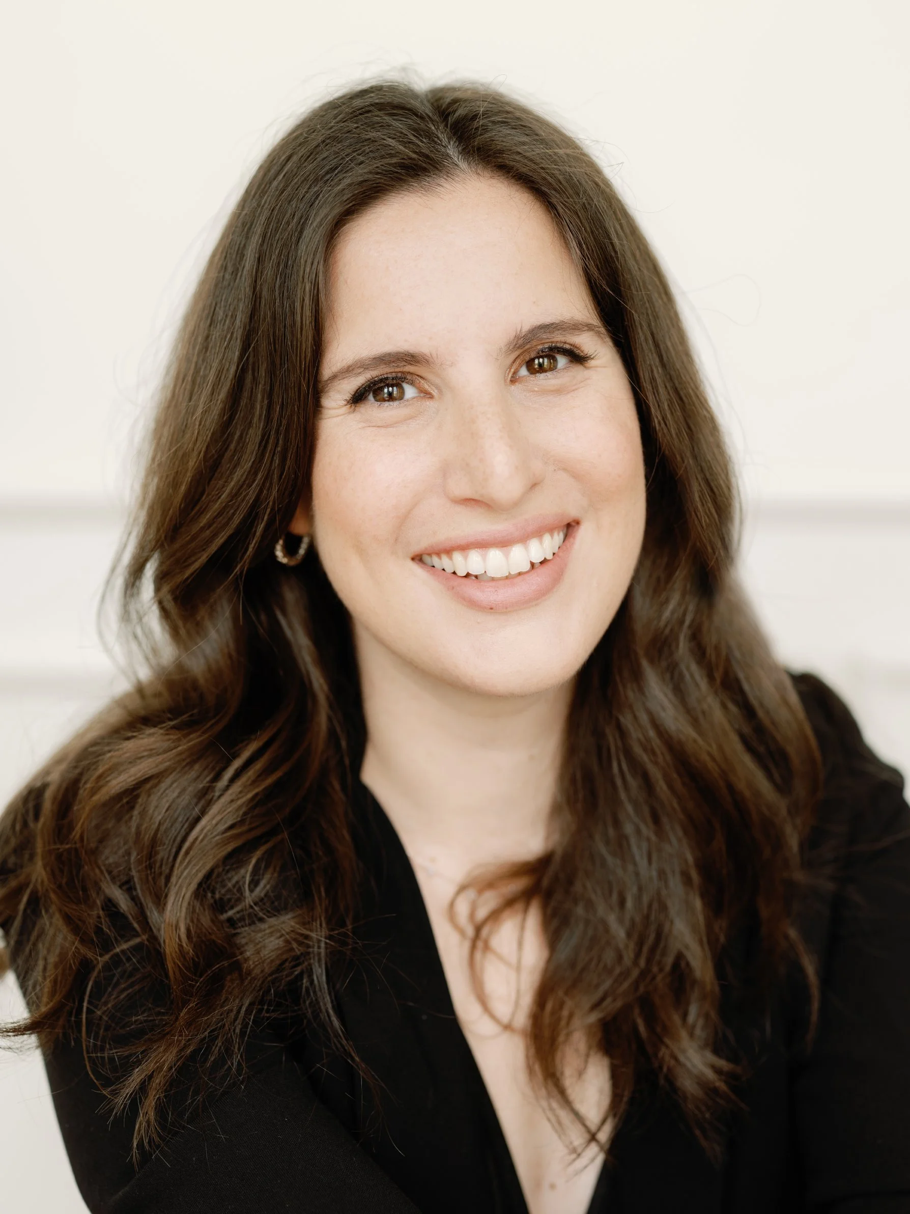Close-up of a woman with long, wavy brown hair, smiling and looking at the camera, wearing a black top and earrings.
