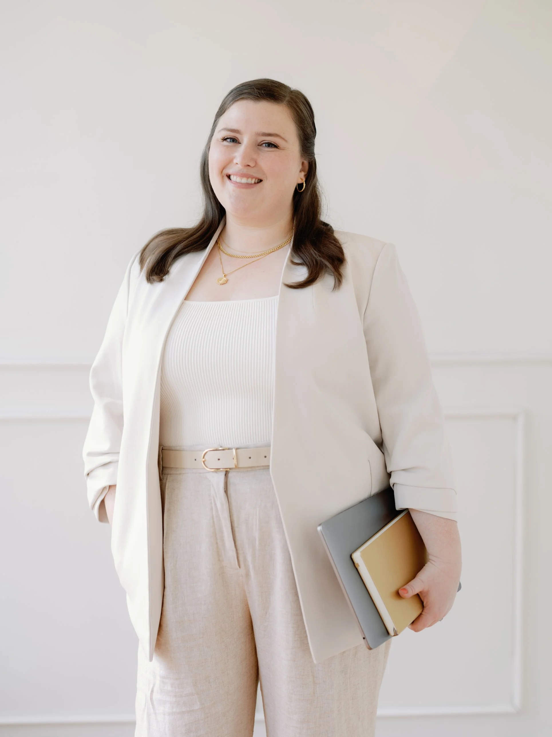 A woman with dark hair wearing a beige blazer, white top, and beige pants, holding notebooks, smiling at the camera against a plain light background.