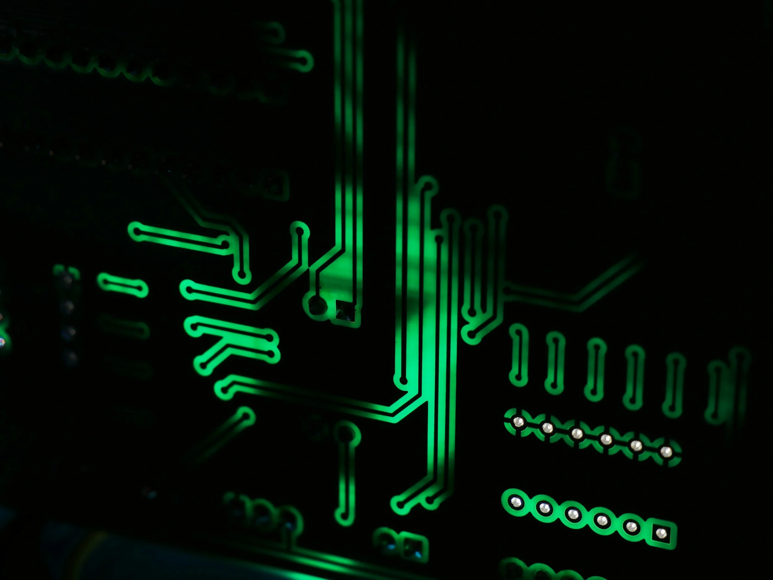 Close-up of a glowing green printed circuit board with various lines and electronic components in a dark background.