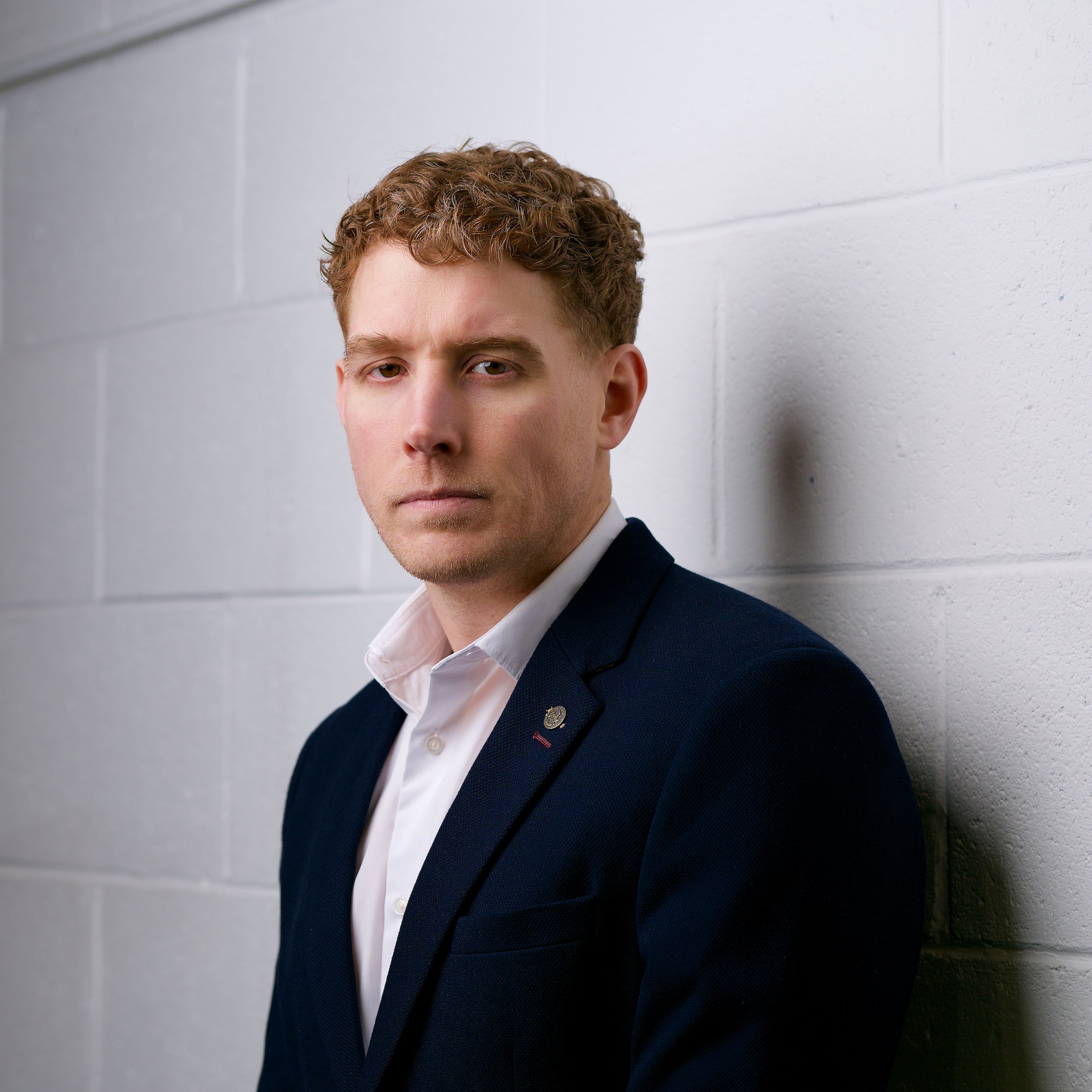 Close-up of a young man with curly reddish-brown hair and fair skin, wearing a dark blazer and white shirt, standing against a white brick wall.