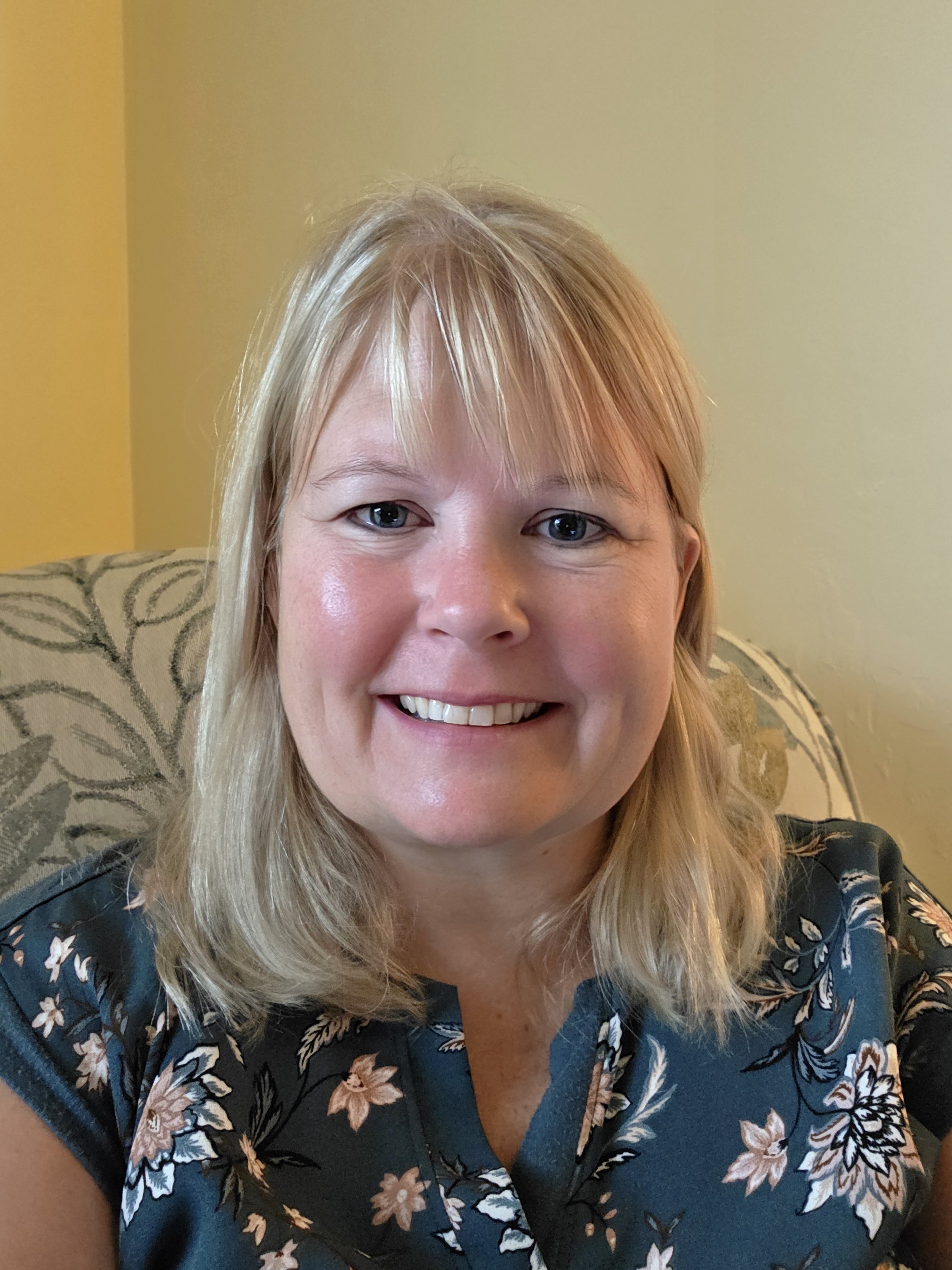 A woman with blonde hair smiling, sitting on a patterned armchair in a room with light yellow walls, wearing a dark floral top.