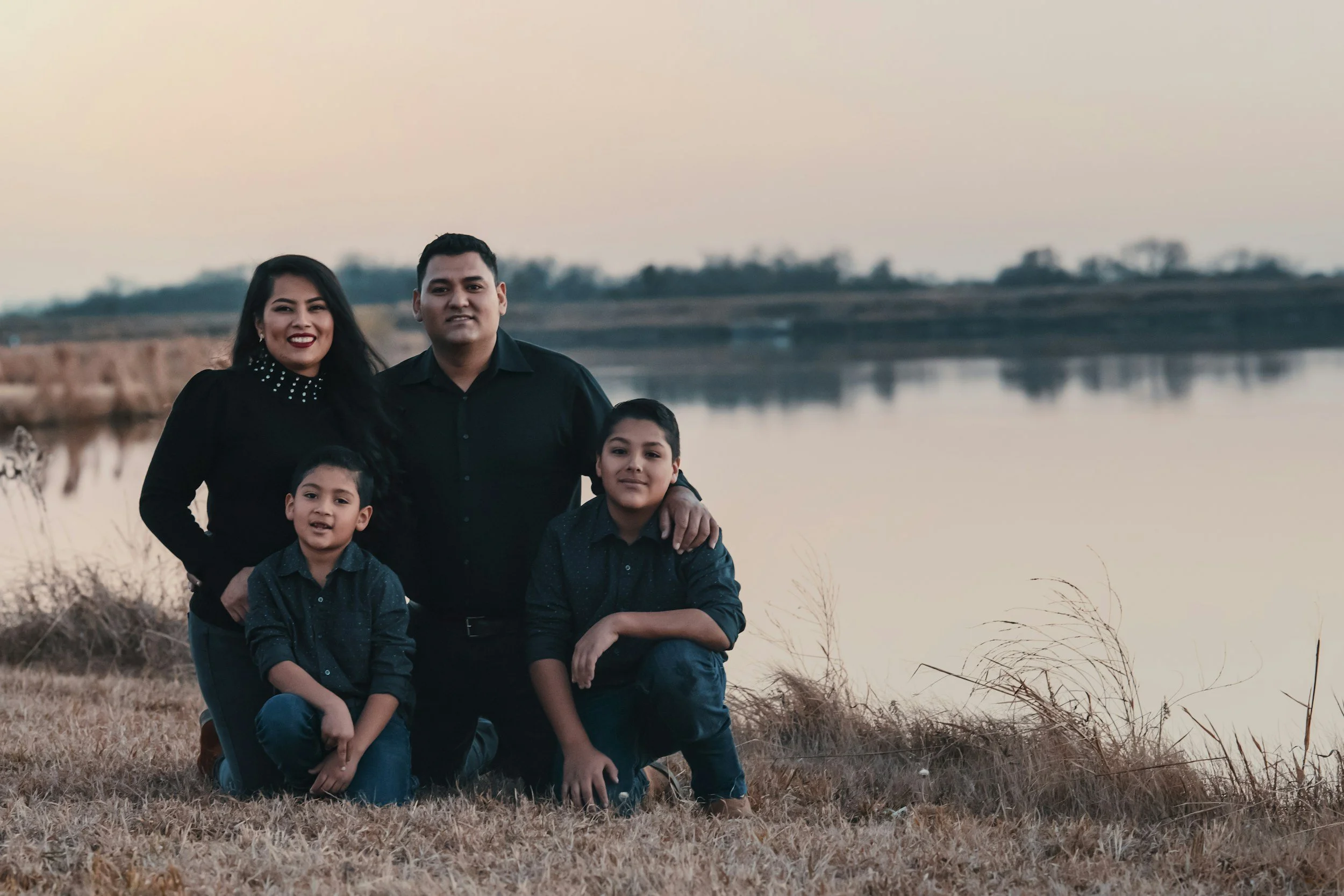 A family of five standing outdoors near a body of water during sunset, with the adults smiling and the children in casual clothing.