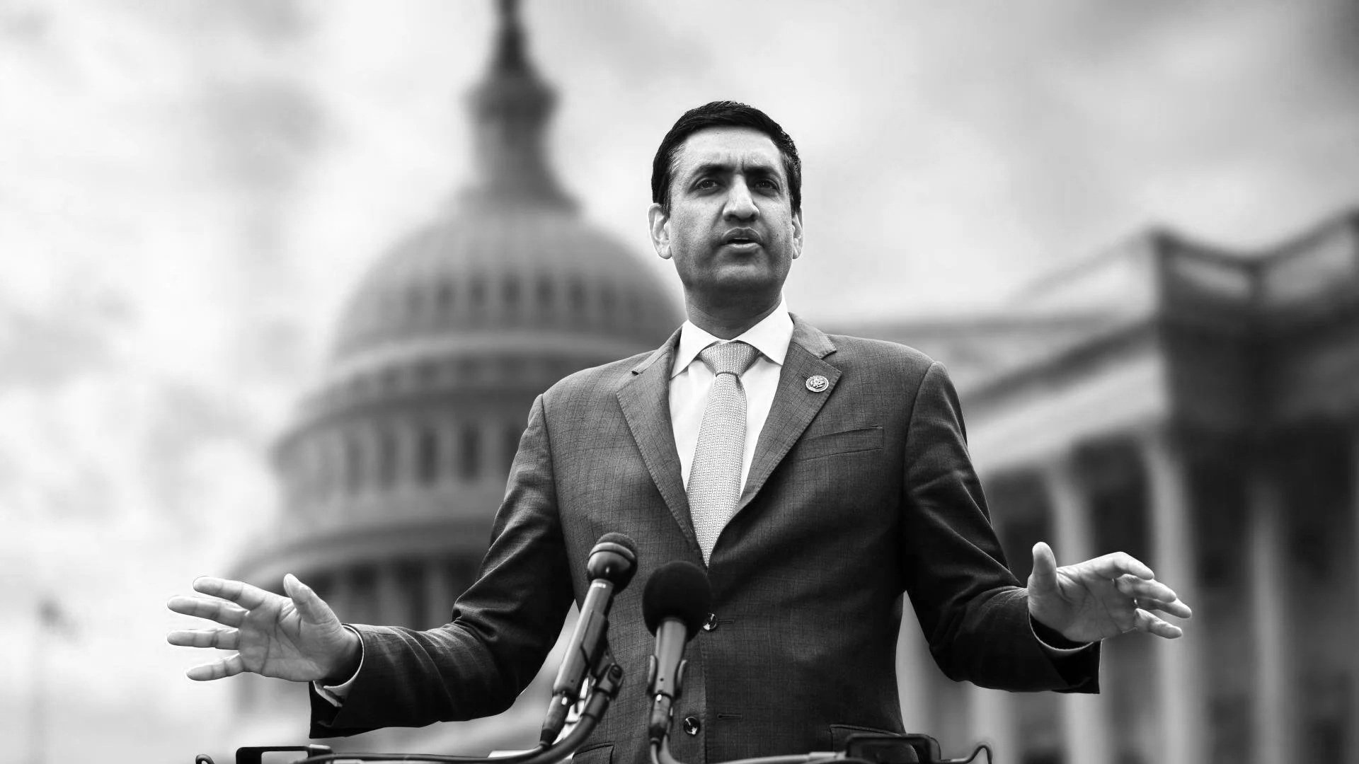 A man in a suit and tie speaking at a podium with microphones, with the U.S. Capitol building in the background, in black and white.