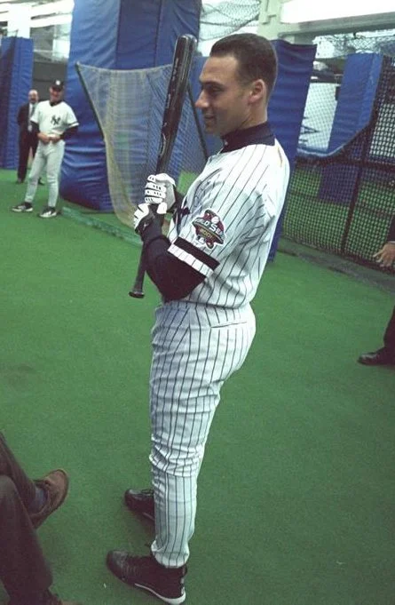 Historical photograph of Derek Jeter in New York Yankees pinstripes holding a bat near a batting cage
