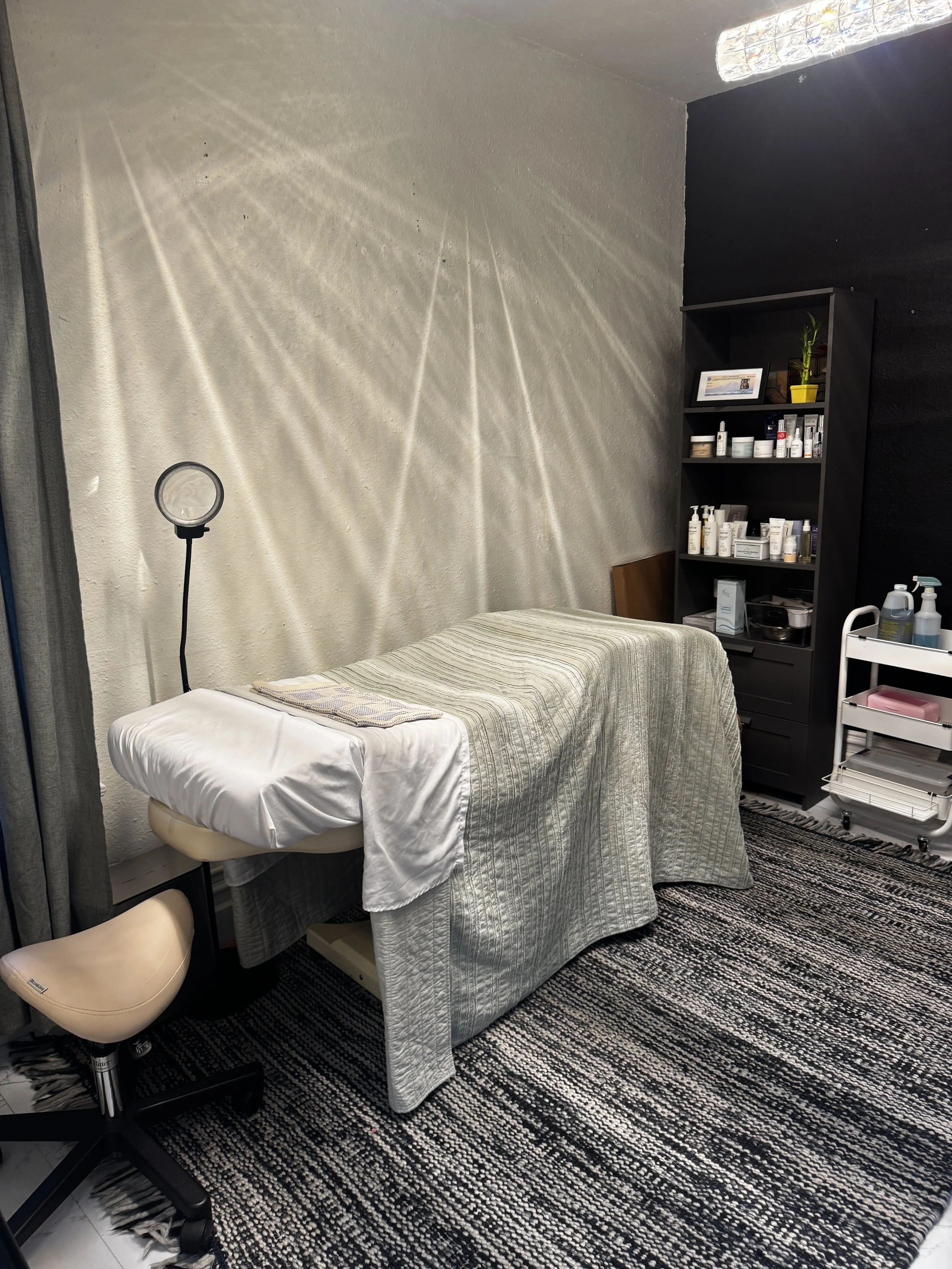 An aesthetic treatment room with a massage bed covered in a striped beige and white cloth. There is a black shelf with skincare products, a framed photo, and a potted plant. A white trolley with bottles and supplies is next to the shelf. The room has a textured black and white rug and walls painted in light and dark tones. A ring light is on the left side of the image, casting light and shadows on the wall.