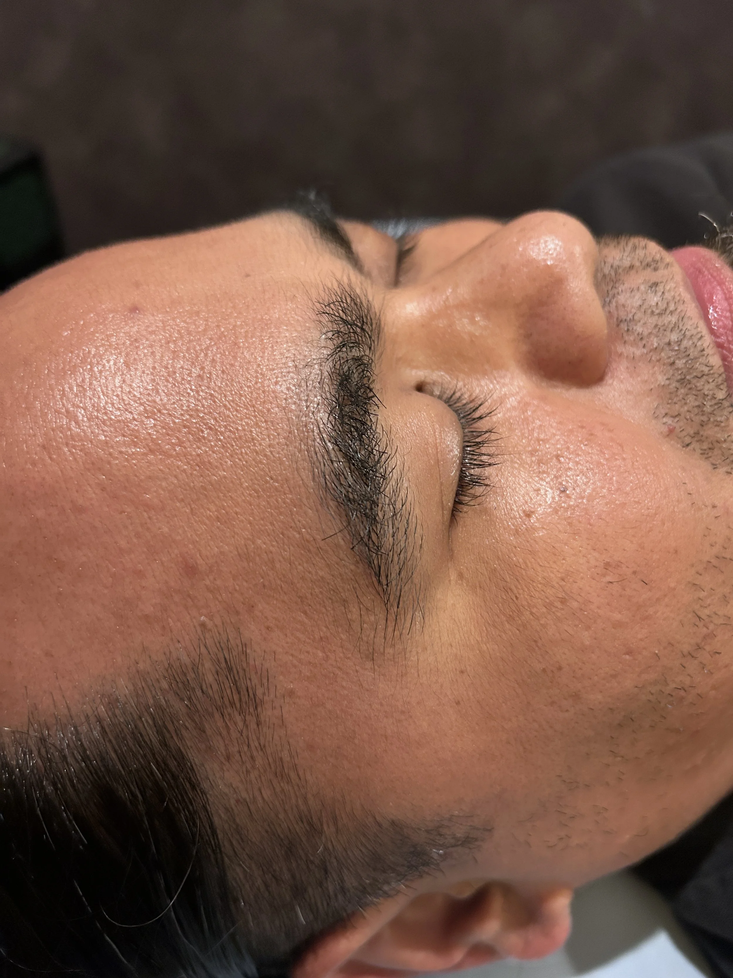 Close-up of a person's face with closed eyes, showing detailed skin and eyelashes, with dark hair and some facial hair visible.