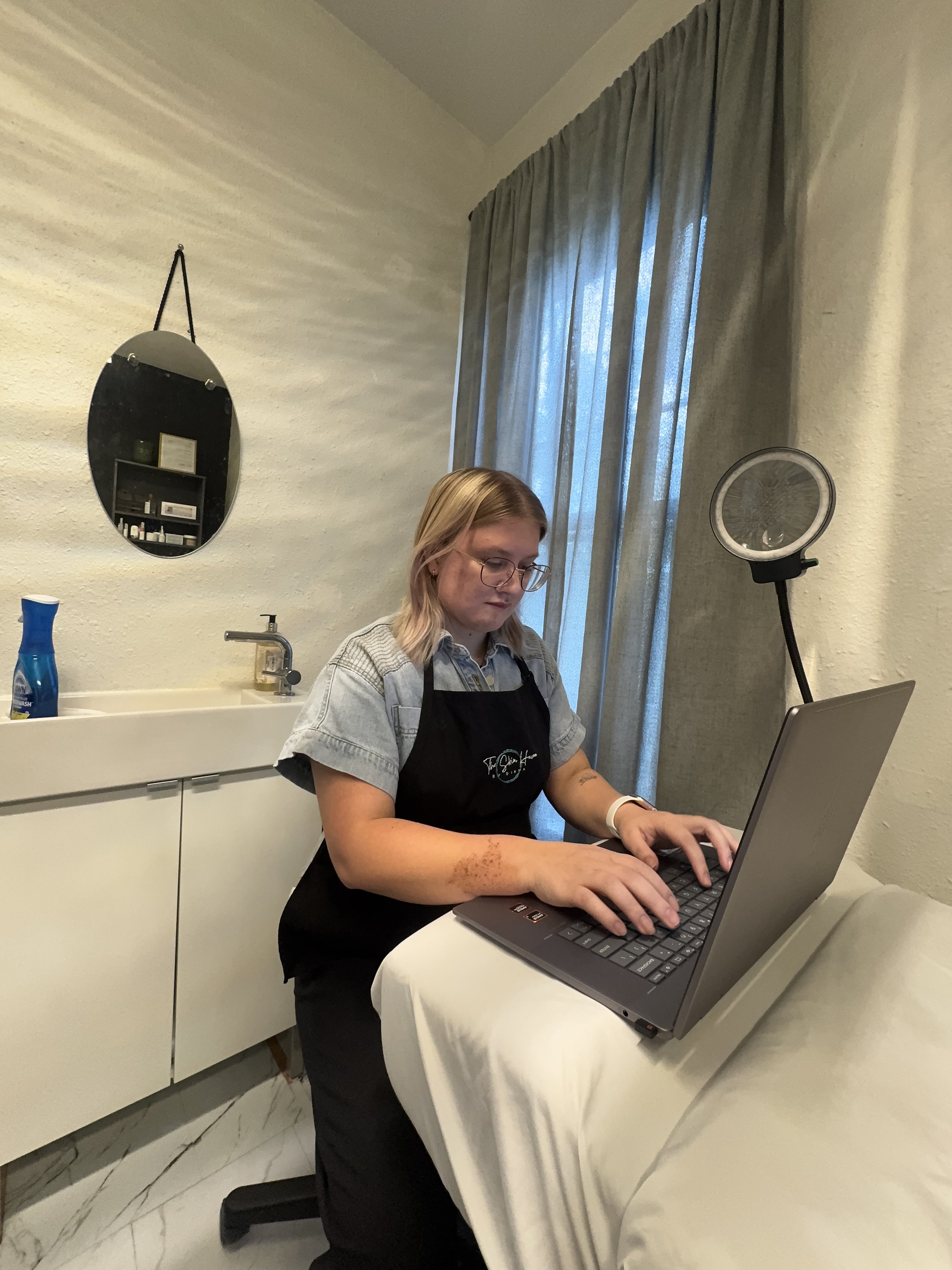 Diana sitting at her esthetics treatment table, working at a computer and wearing an apron
