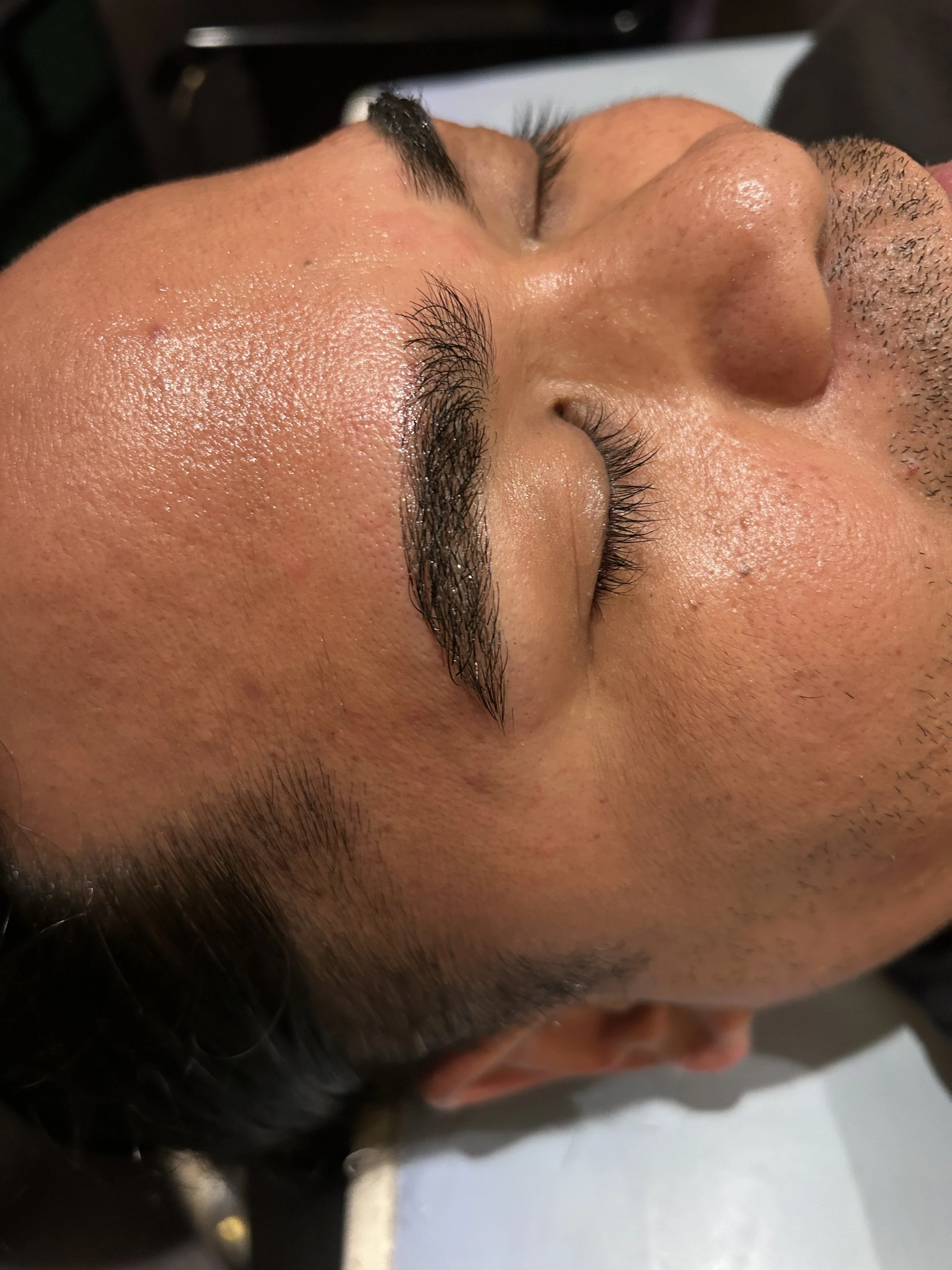 Side view of a person's face with closed eyes and facial hair, possibly receiving a skincare or cosmetic treatment.