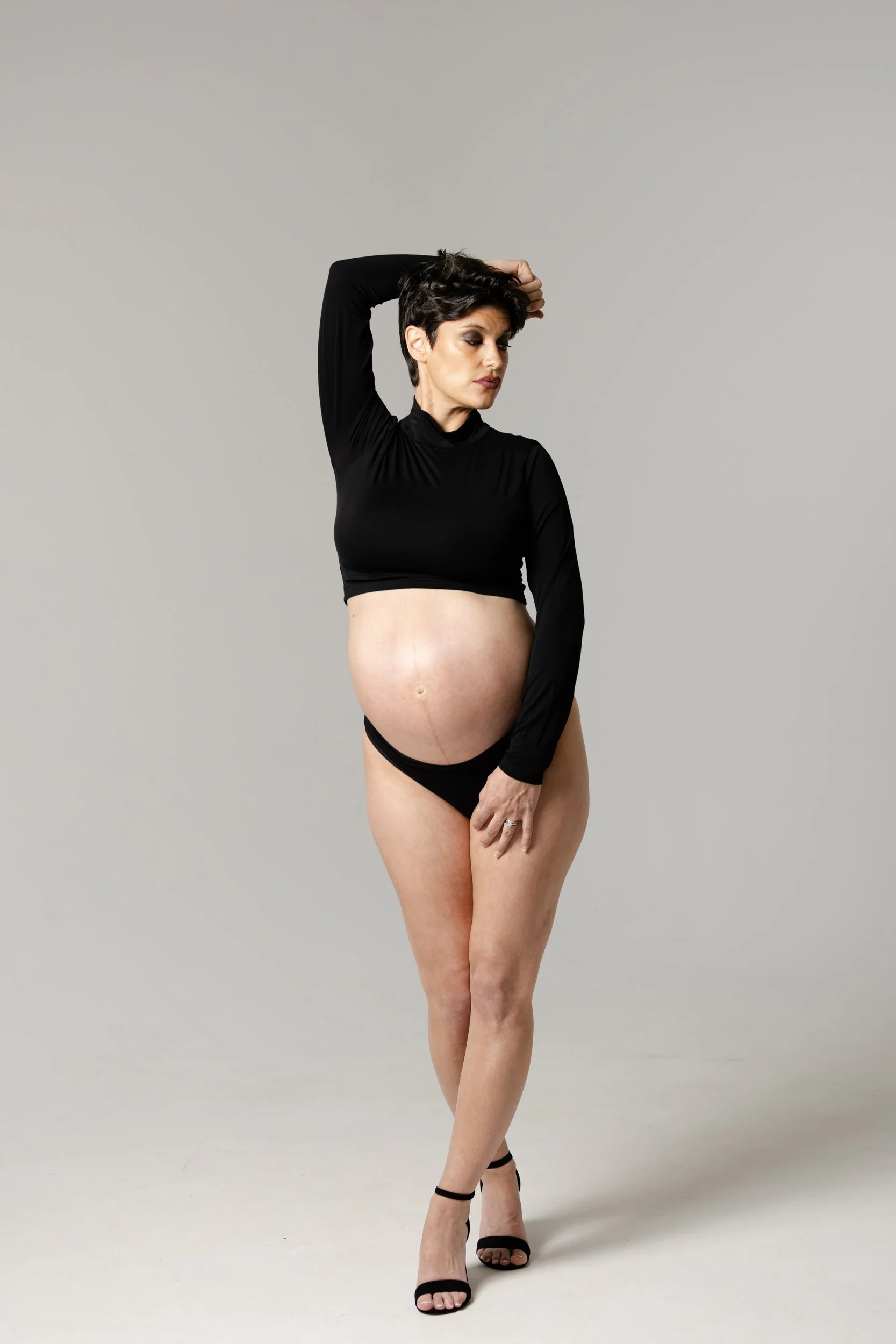 A woman with short dark hair wearing a black long-sleeve crop top, black underwear, and black high-heeled sandals standing with one leg crossed over the other against a neutral background.