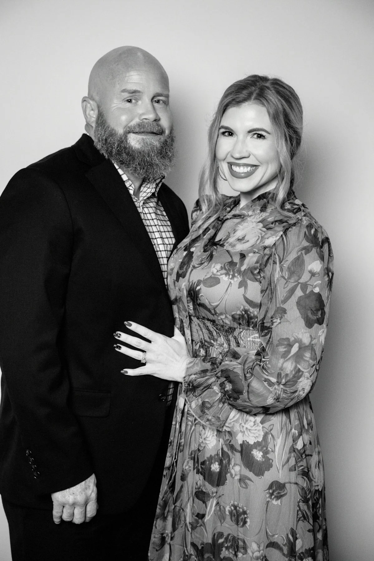 A black and white photo of a smiling man with a bald head and beard wearing a dark suit, and a smiling woman with wavy hair wearing a floral dress, standing close together against a plain background.