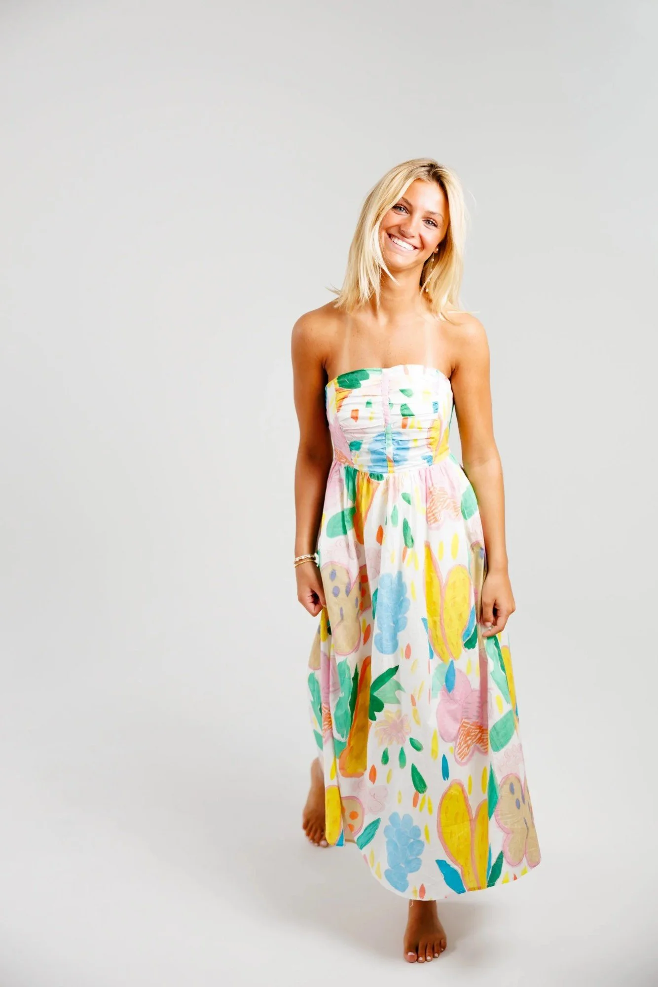 A smiling woman with blonde hair standing barefoot in a colorful, flowy, strapless dress with a floral and abstract pattern against a plain light background.