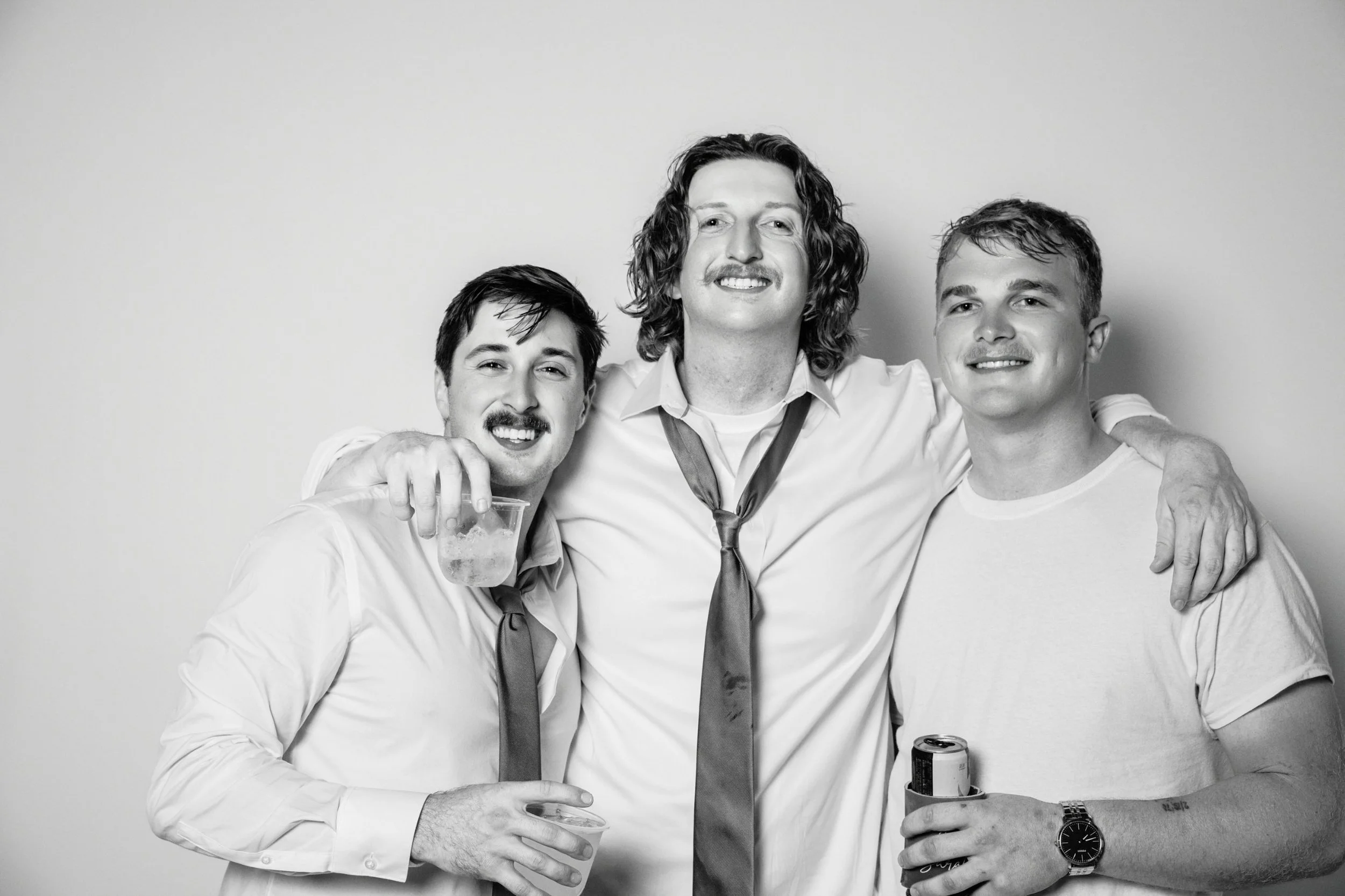 Three men celebrating, smiling, with one holding a drink and another holding a can. They are dressed casually, with one wearing a watch, and they have their arms around each other against a plain background.