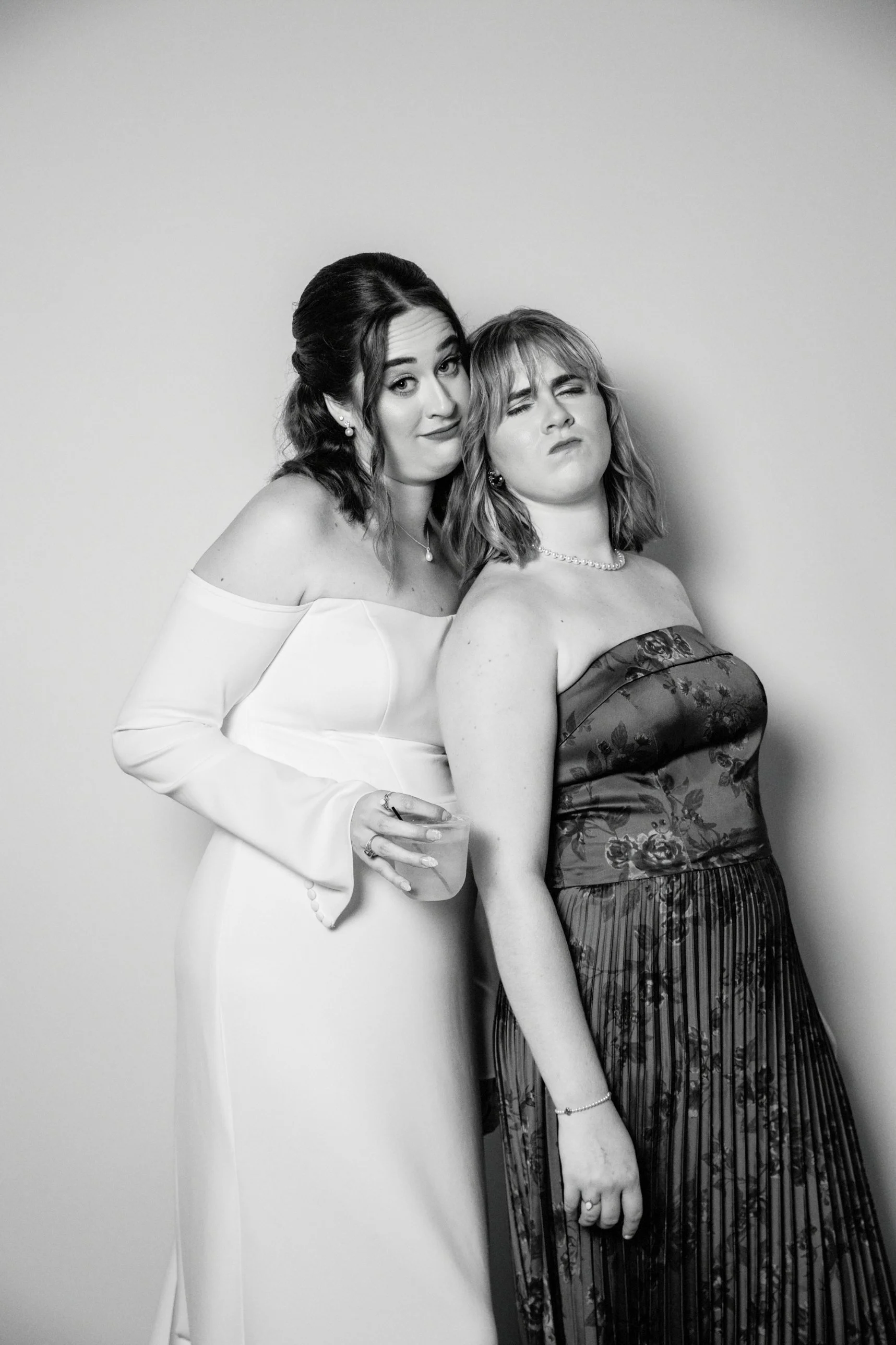 Two women dressed elegantly, with one in a white off-shoulder gown holding a drink, and the other in a floral strapless dress, posing against a plain wall.