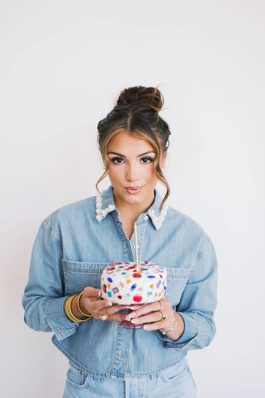 Young woman with styled hair, wearing denim jacket with pearl details, holding a colorful birthday cake with a single lit candle, against a plain white background.