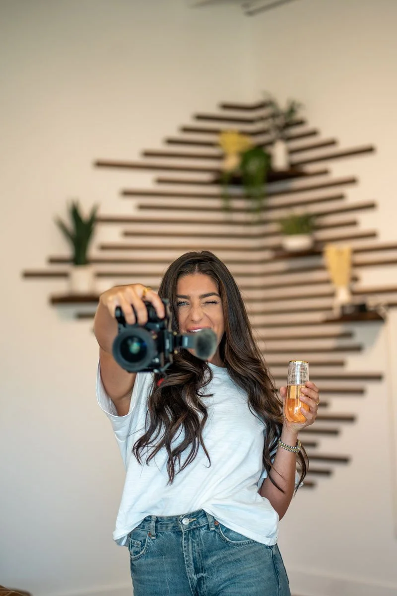 A woman with long dark hair takes a selfie with a camera while holding a drink in a modern indoor setting with a decorative wooden wall and plants in the background.