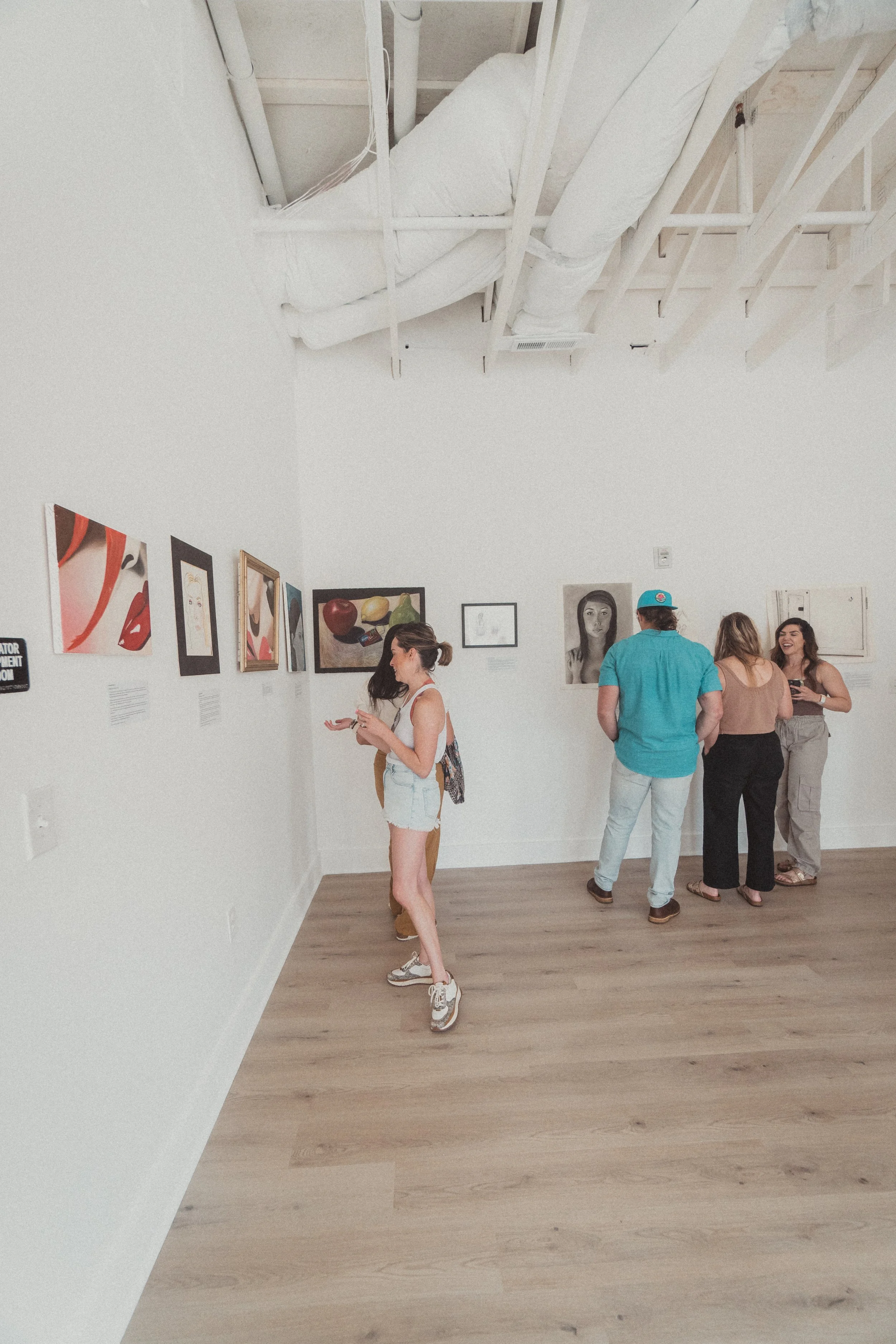 People viewing artwork at an art gallery, with paintings hanging on white walls and a light wood floor.