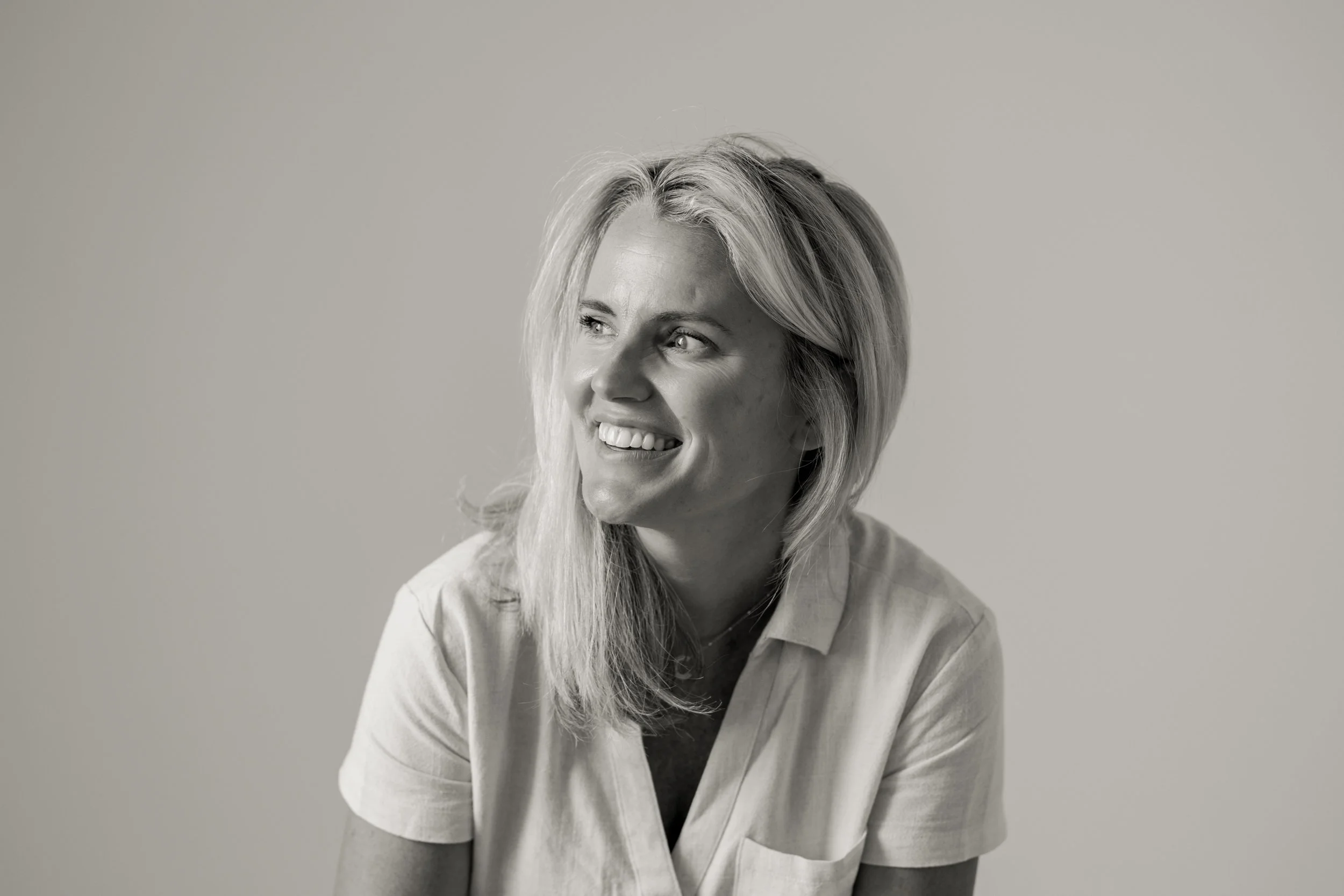 A woman with long blonde hair smiling and looking to her right, wearing a light-colored shirt, in front of a plain background.