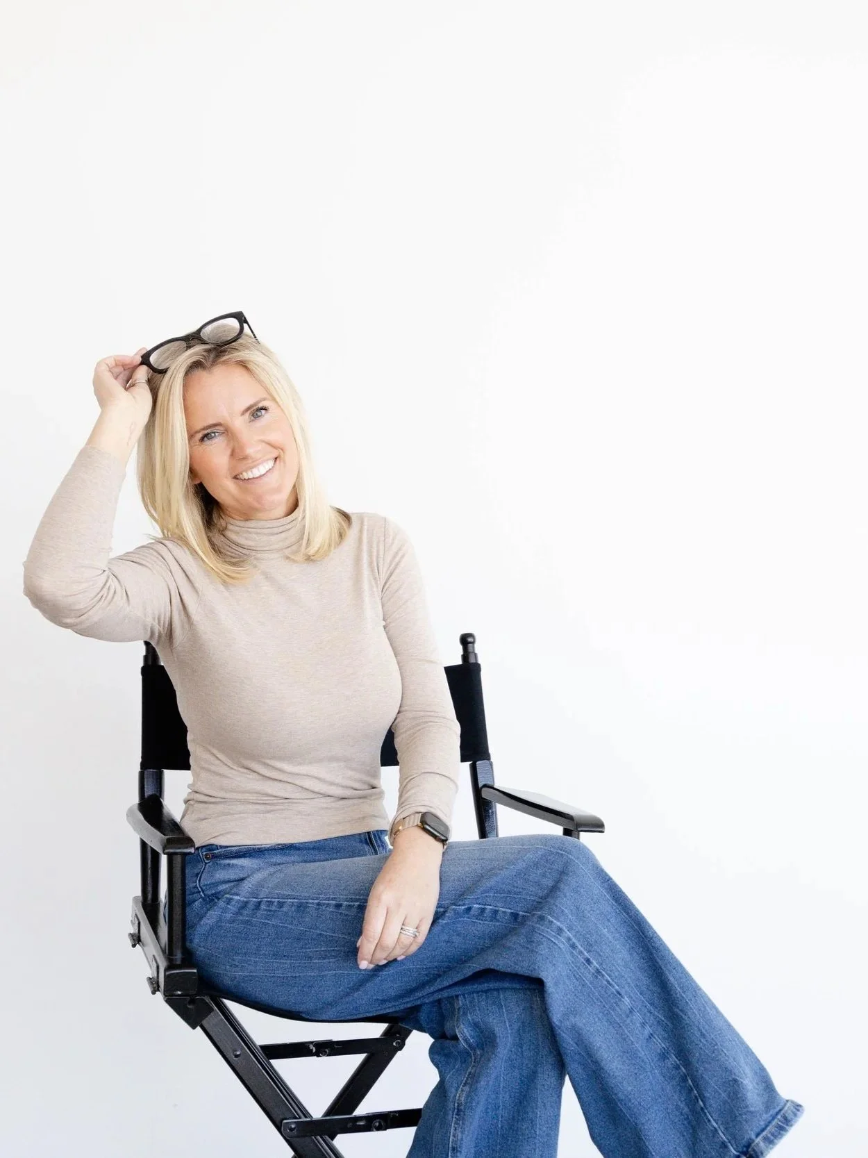 Woman sitting on a director's chair, smiling, holding glasses on her head, wearing a beige turtleneck and blue jeans, against a white background.