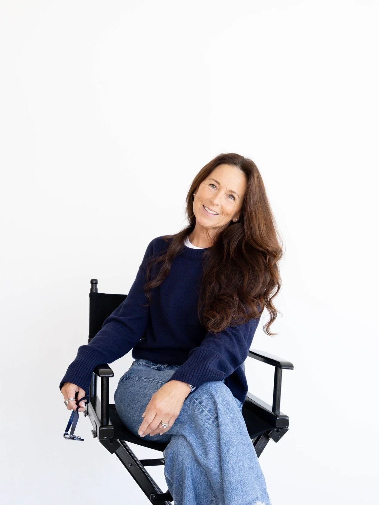 A woman sitting on a director's chair, smiling, with long wavy brown hair, wearing a navy blue sweater and light blue jeans, against a plain white background.