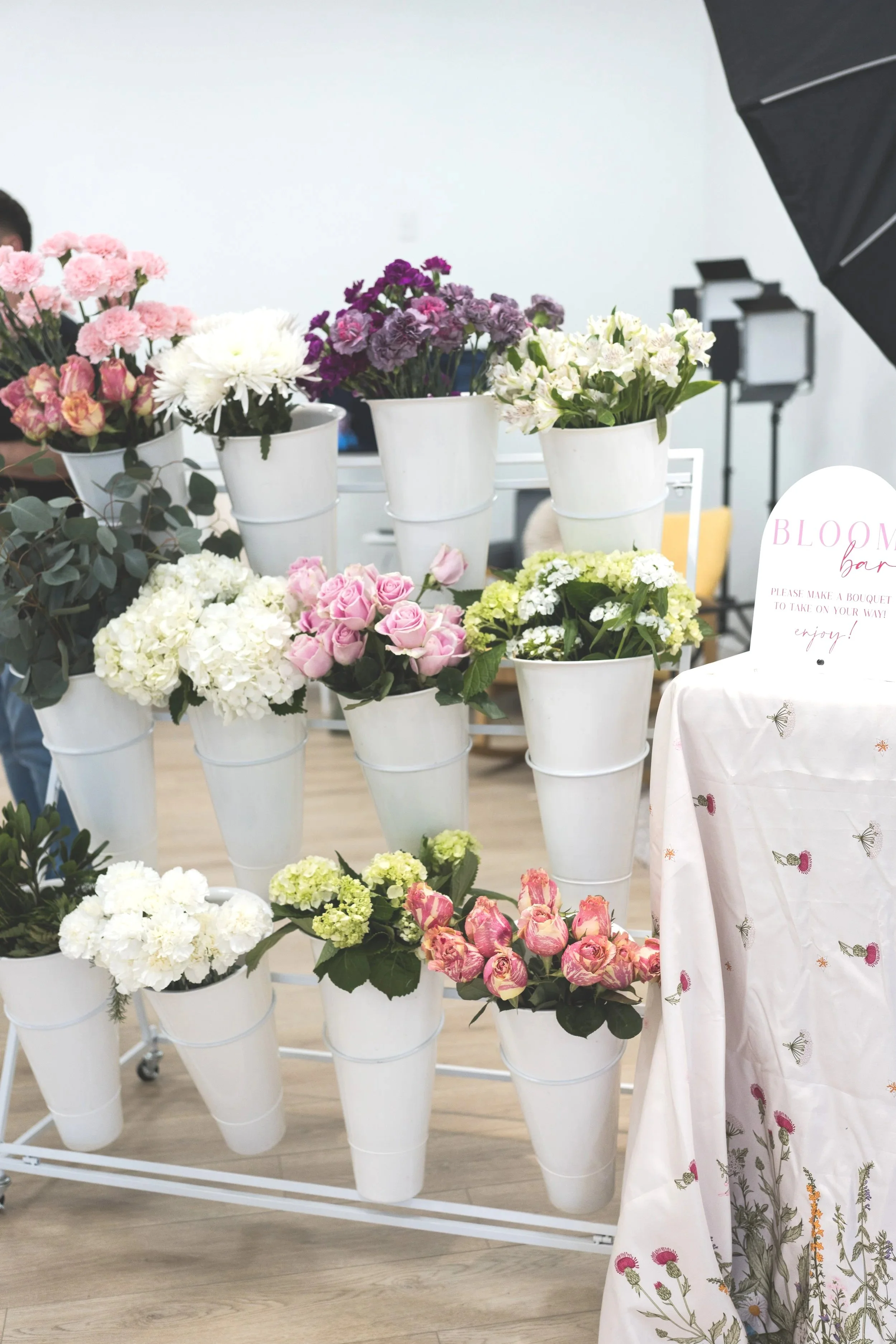 Various colorful flowers arranged in white buckets on a white mobile stand, with a backdrop of a photography studio and a table with a floral tablecloth.